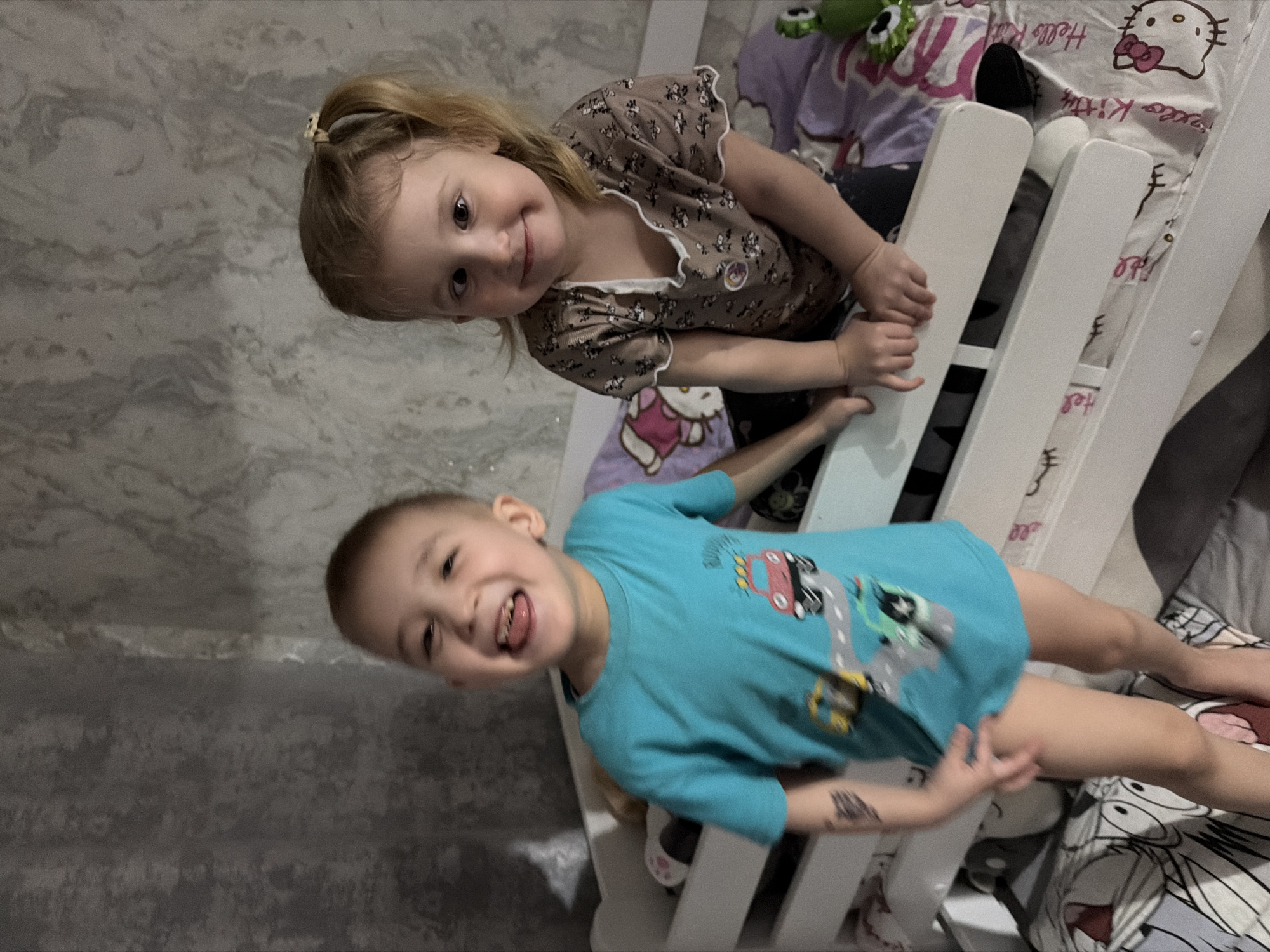 Two children, a girl and a boy, are positioned on a white bunk bed with a Hello Kitty-themed blanket. The girl is wearing a brown floral blouse, while the boy wears a blue t-shirt with car graphics and is sticking out his tongue. The backdrop consists of a textured gray wall. The location or presence of Noviy, Russia, is not discernible from the image.