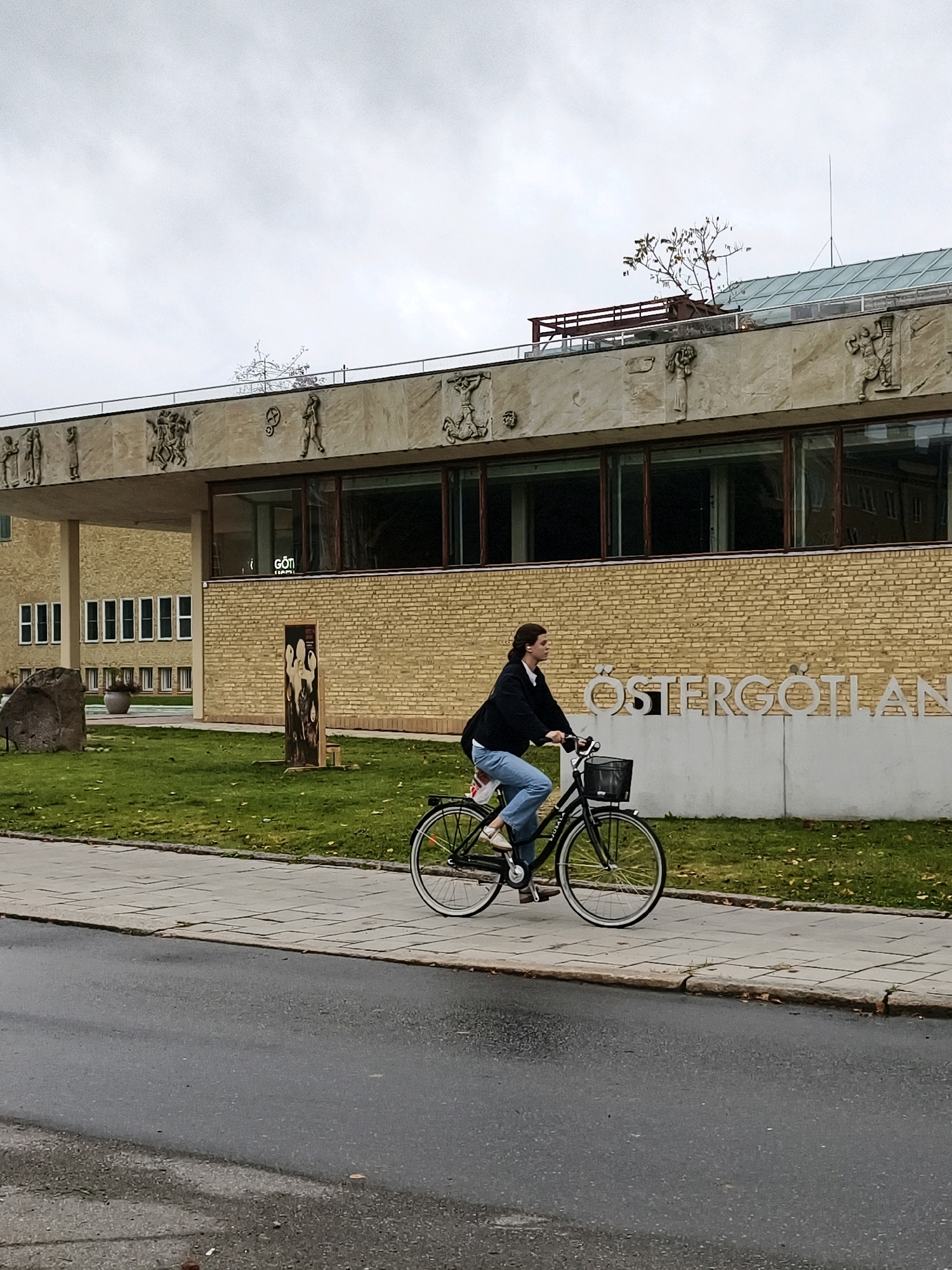 A young woman is riding a black bicycle with a basket on the handlebars down a paved sidewalk. She is wearing a dark jacket, a white shirt, and blue jeans. The bicycle has a white stripe on the tire. The sidewalk is lined with grass and trees. In the background, there is a building with light-colored brick and stone facade. The building has large windows and decorative sculptures above them. A sign on the building reads "ÖSTERGÖTLAND". The scene is likely in Linköping, Sweden, given the presence of the "ÖSTERGÖTLAND" sign, as Östergötland is a historical province and county in Sweden, and Linköping is its capital. The sky is overcast.