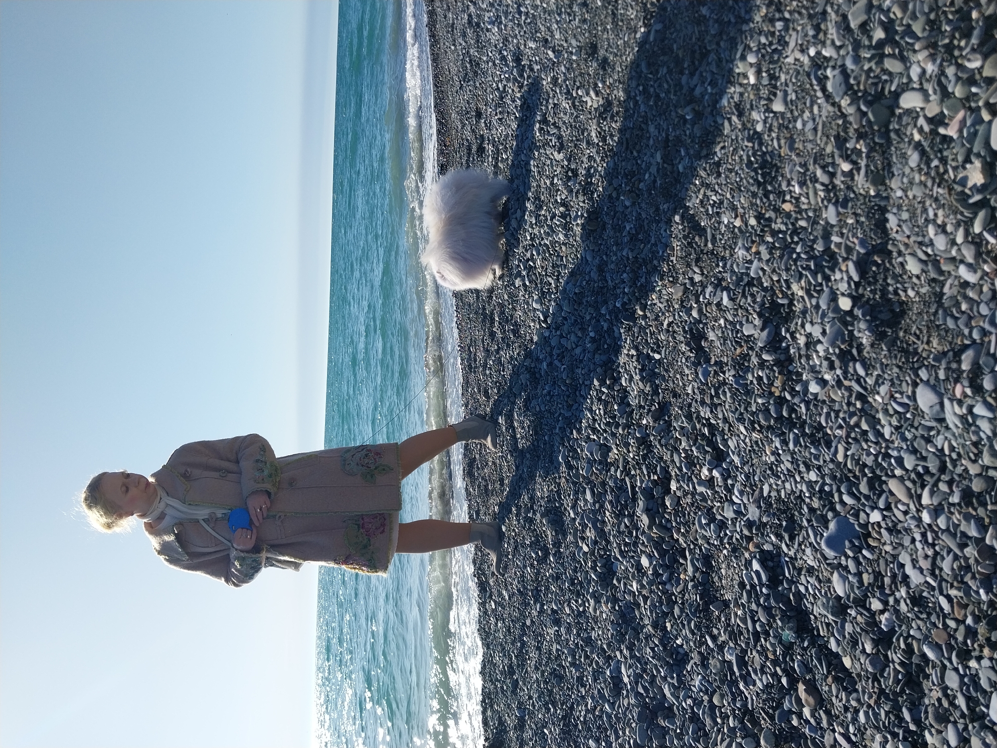 The image depicts a tranquil coastal scene on a pebble beach in Sochi, Russia. A woman and a fluffy white dog are the central subjects, positioned along the shoreline under a clear blue sky.
The woman stands on the dark-toned pebble beach, facing towards the calm, turquoise sea. She has blonde hair pulled back and is dressed in a long, beige-colored coat with intricate embroidered or patterned details, a light scarf, and boots. She holds a small, bright blue object in her hands. Distinct shadows of both the woman and the dog are cast on the pebbled ground, indicating bright sunshine and suggesting either mid-morning or late afternoon.
To the woman's right, and slightly closer to the water, is a small, white, very fluffy dog, appearing to be observing the gentle waves as they reach the shore. The sea exhibits a beautiful range of blues and greens, with sunlight sparkling on its surface. The beach consists entirely of smooth, rounded pebbles of varying sizes, predominantly in shades of gray, black, and brown. There is no visible text in the media file.