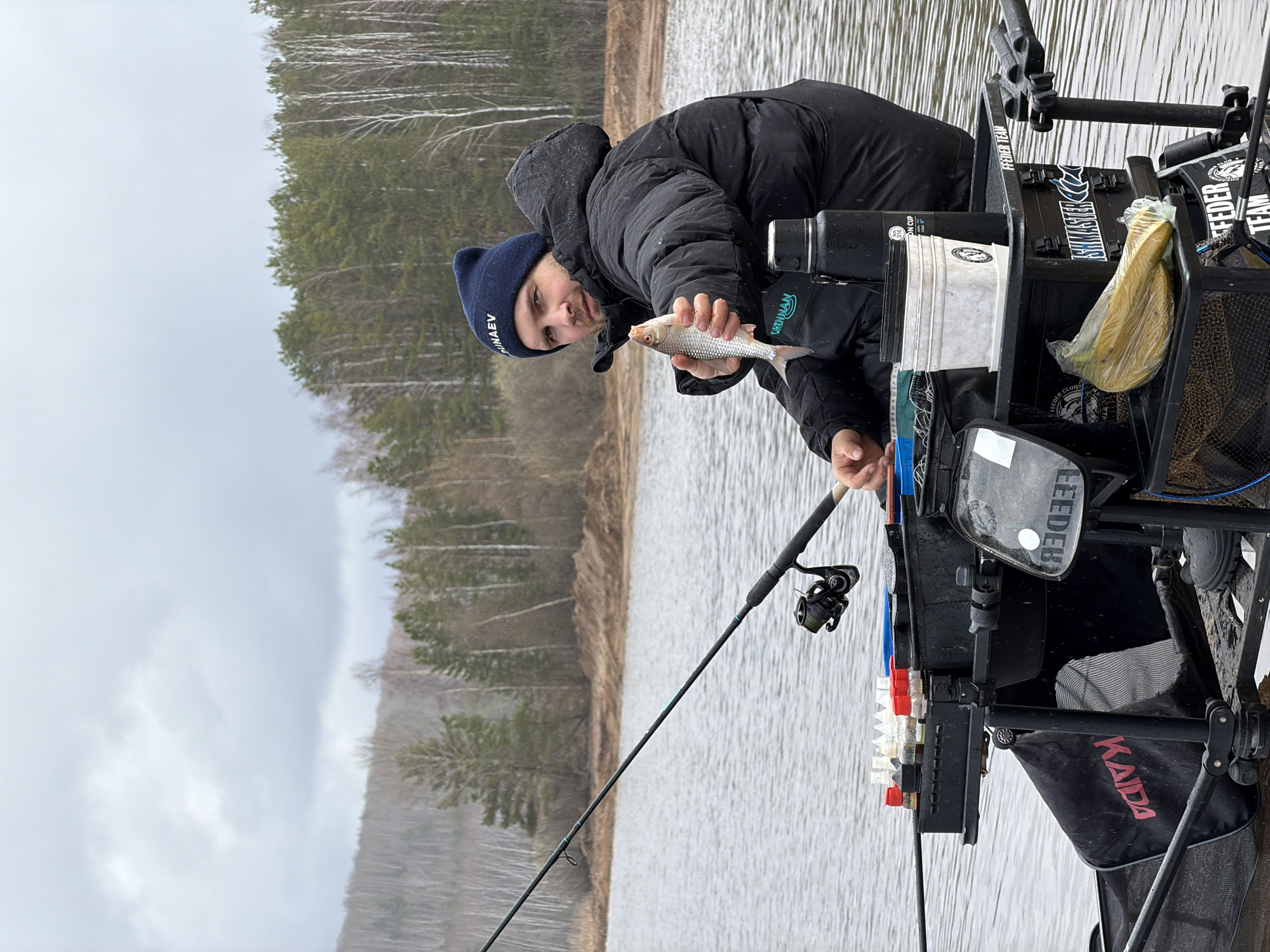 A man in a dark blue beanie and a dark winter coat stands at the water's edge, holding a small fish in his hand. He is positioned near a complex fishing setup, which includes a black chair or platform with various attachments and containers for fishing gear. A fishing rod is visible, extending out over the water. The background consists of a body of water, possibly a lake or wide river, with a line of bare trees visible on the opposite shore. The sky is overcast, suggesting a cool or cold day. The man is looking at the camera, seemingly showing off the fish he caught. Text on the beanie appears to read "ПYHA...EV." On the fishing setup, the words "Feeder Team" are partially visible on a logo or sign. Another sign says "Fishmaster". The location is likely near a body of water in the city of Chaykovsky, Russia.