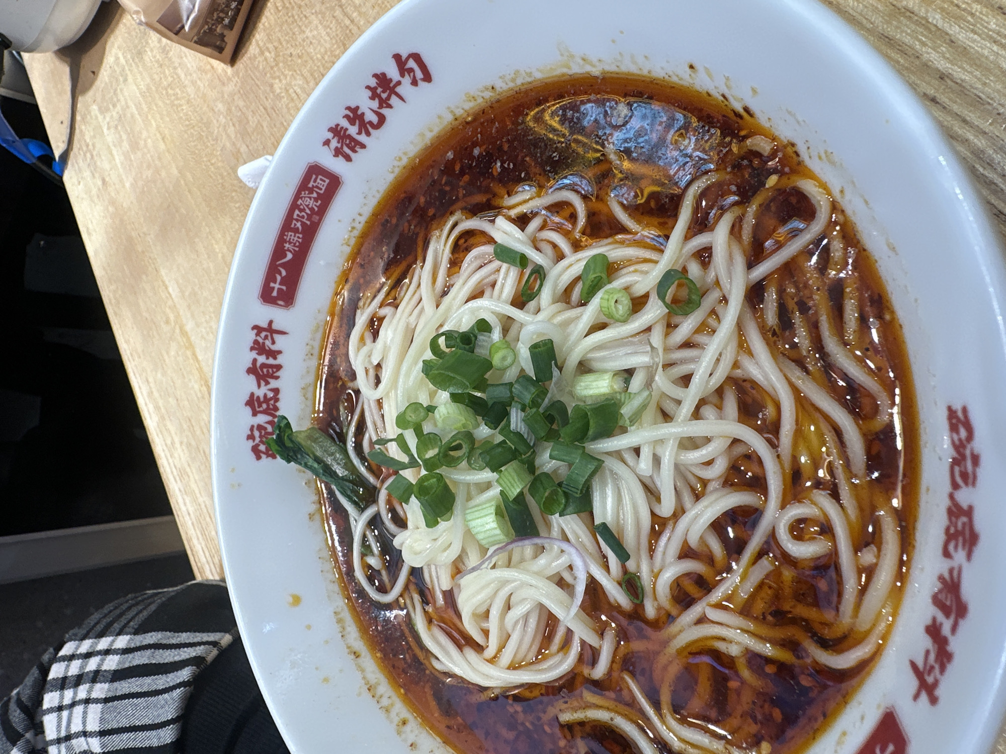 This close-up, high-angle photograph captures a bowl of spicy Chinese noodles placed on a light-colored wooden table. The white ceramic bowl contains thin, white noodles immersed in a rich, reddish-brown chili oil-based sauce or soup, typical of Chongqing cuisine. The noodles are generously garnished with chopped green onions and a single green leafy vegetable. The soup exhibits an oily sheen with visible chili flakes and spices.
Red Chinese characters adorn the rim of the white bowl. Part of the visible text reads "请先拌匀" (Qǐng xiān bàn yún), which translates to "Please mix well first," suggesting an instruction for consumption. A red rectangular label within the bowl's design prominently displays "十八梯邓矮面" (Shíbātī Dèng'ǎi Miàn), identifying the dish or establishment as "Shibati Deng'ai Noodles." Another phrase, "有料" (Yǒuliào), meaning "has ingredients" or "is substantial," is also visible.
In the bottom left corner, a black and white checkered pattern, likely from a sleeve or jacket, indicates the presence of a person seated at the table. The wooden table surface extends across the upper left of the frame, with a blurry brown package or item also visible. The setting is identified as Chongqing High-tech Zone, China.