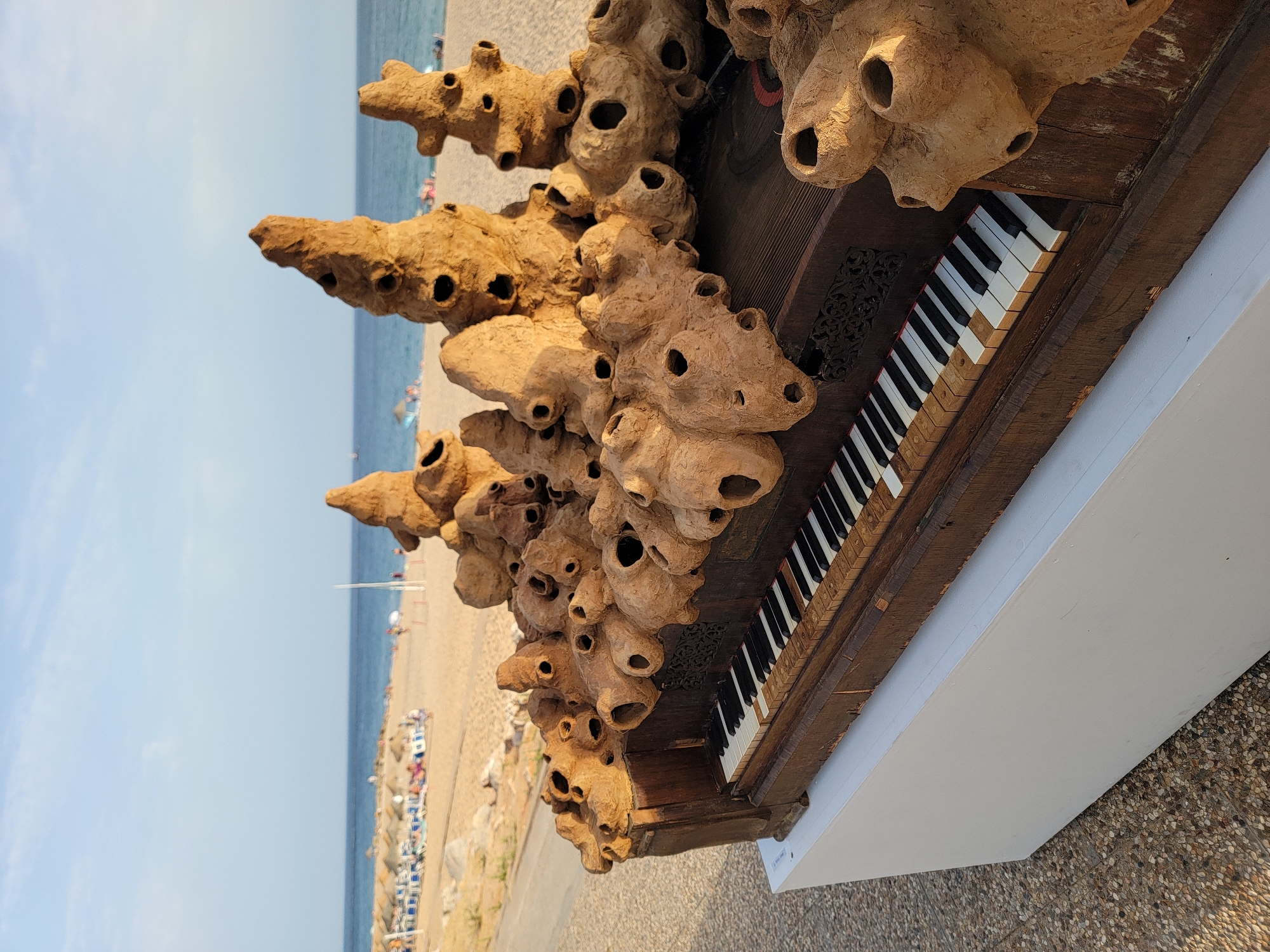 A sculpture resembling a cluster of organic, porous formations is placed on top of an antique piano. The piano has visible wooden keys, some of which are discolored or chipped. The sculpture is made of a rough, textured material, possibly clay or a similar substance, and features numerous holes of varying sizes. The piano and sculpture are displayed outdoors on a white pedestal.
In the background, a sandy beach with beach umbrellas and lounge chairs is visible. People are scattered on the beach and in the shallow water. The sea stretches to the horizon under a bright, clear sky with a few wispy clouds. The location appears to be a coastal area, likely a beach resort. This scene is located in the city of Quarteira, Portugal, a popular tourist destination known for its beaches.