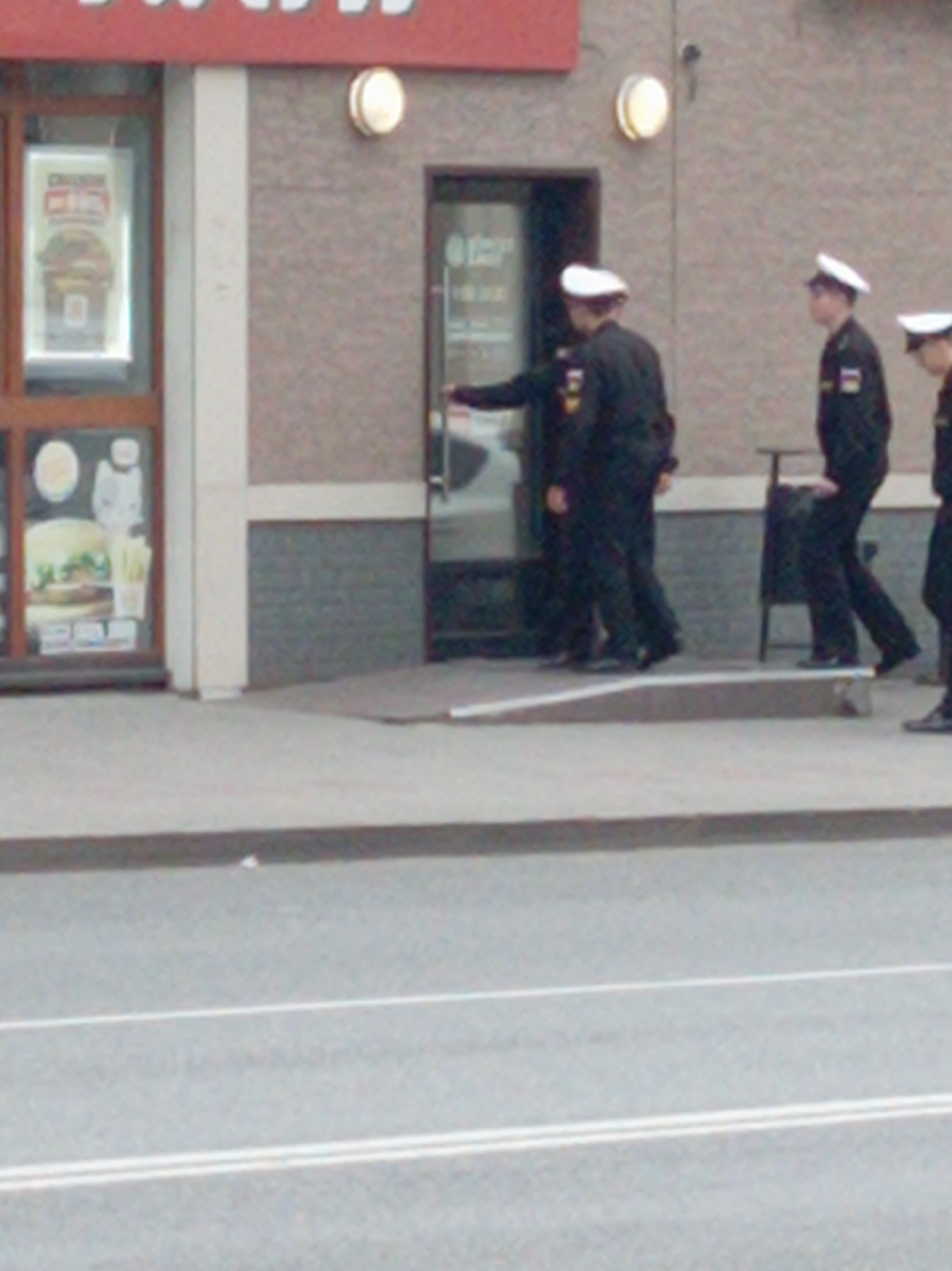 The image captures a street scene in Vladivostok, Russia. In the background, a building with a red awning is visible. A doorway with a glass door and what appears to be a restaurant sign inside is situated to the right of the building's entrance. Several individuals in dark uniforms and white caps are walking. The individuals are positioned on a paved surface. The foreground shows a street with white lines painted on it.