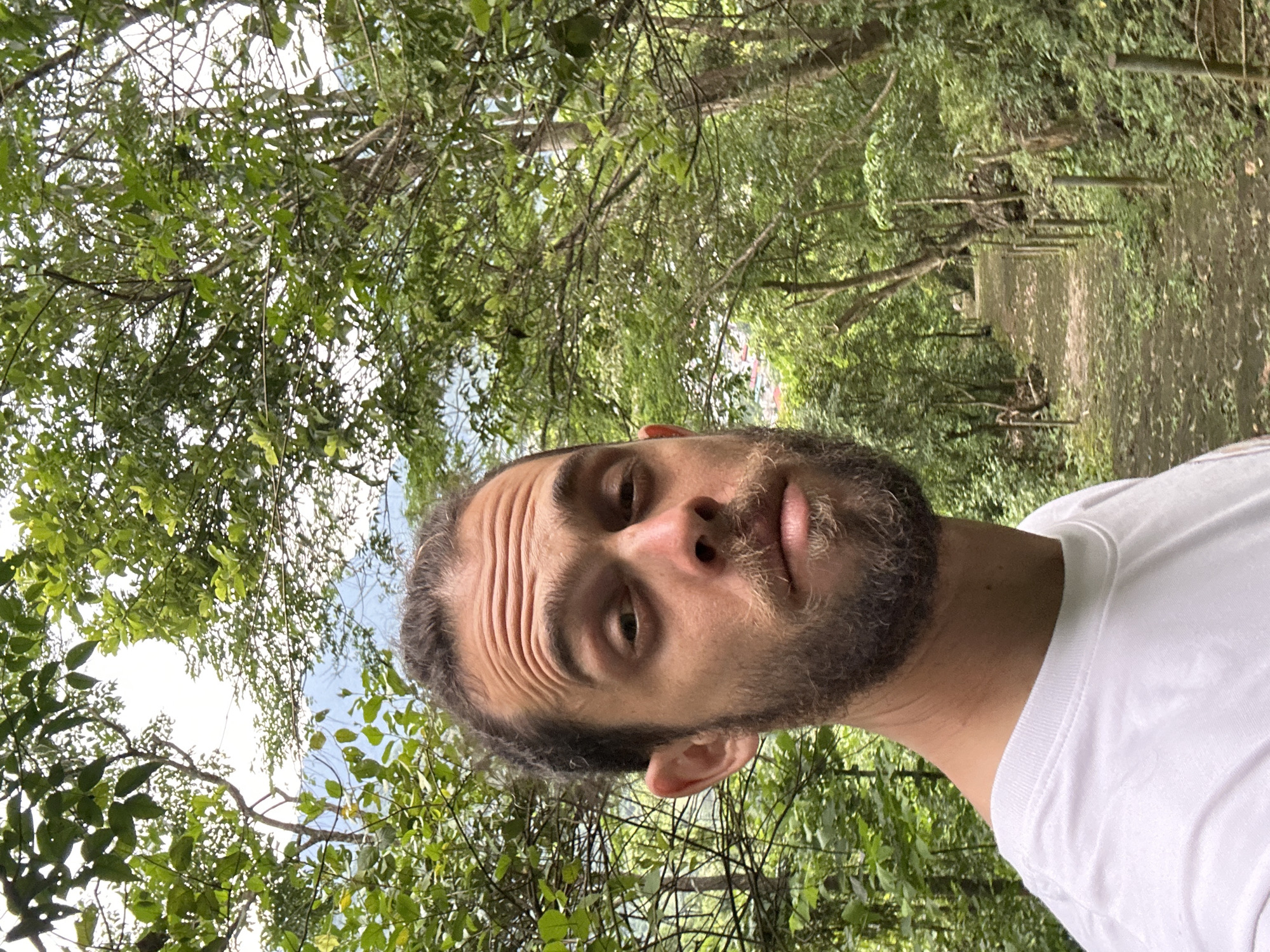 A man with a beard and hair tied back in a bun, wearing a white t-shirt, takes a selfie in a forest. He is positioned in the foreground, with trees and foliage surrounding him. A path with wooden posts lining its sides recedes into the background, indicating a natural outdoor setting. The sky is visible through the dense canopy of leaves. The location is identified as being in the city of Mai Chau, Vietnam.