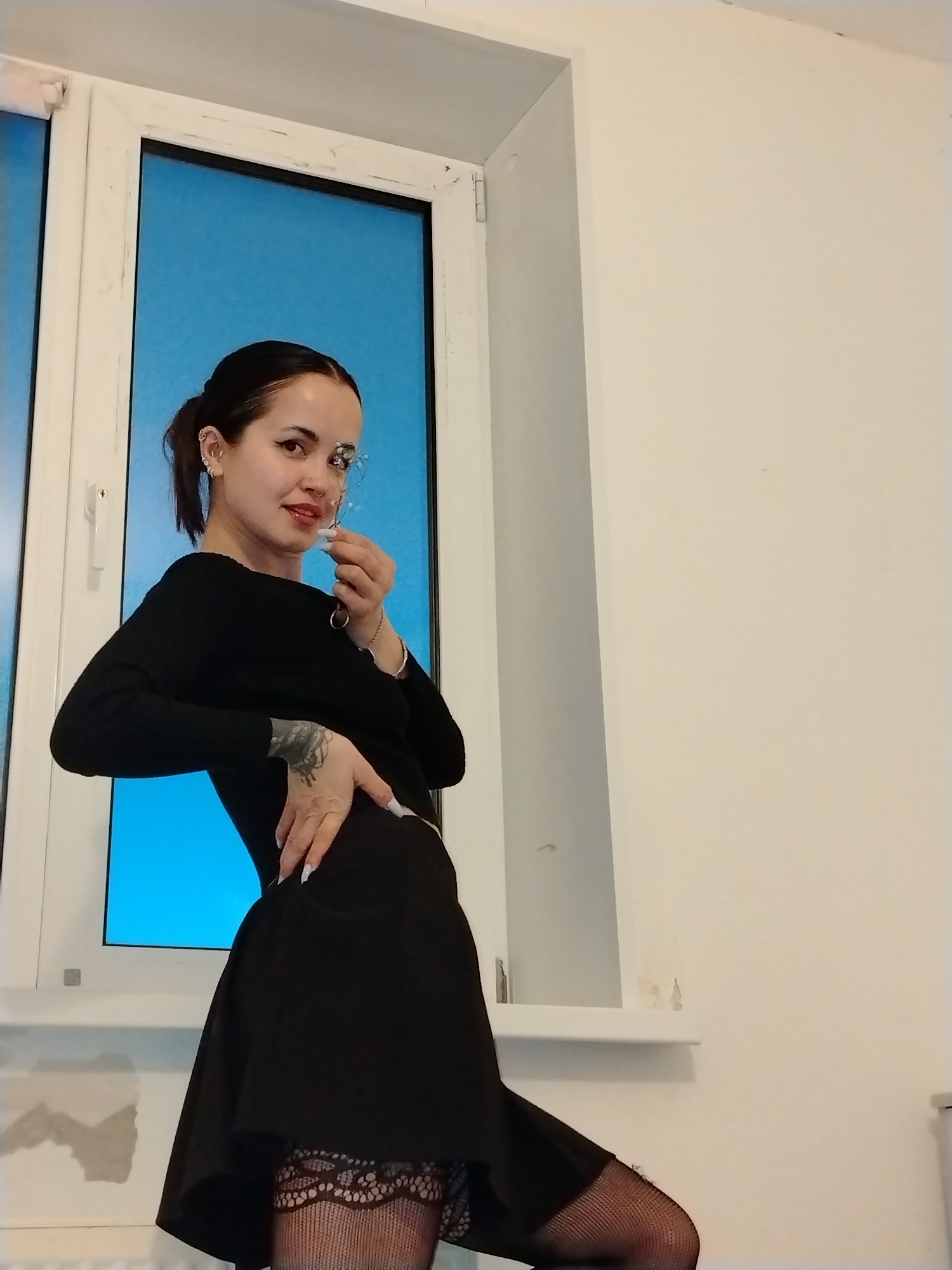 A woman stands indoors next to a window. She is wearing a long-sleeved black top, a short black skirt, and patterned black stockings. She is holding a small object in her right hand near her face. Her hair is pulled back. The window behind her has a white frame and appears to look out on a blue surface. The wall beside the window is plain white. There is no evidence to indicate the image was taken in Noviy, Russia.