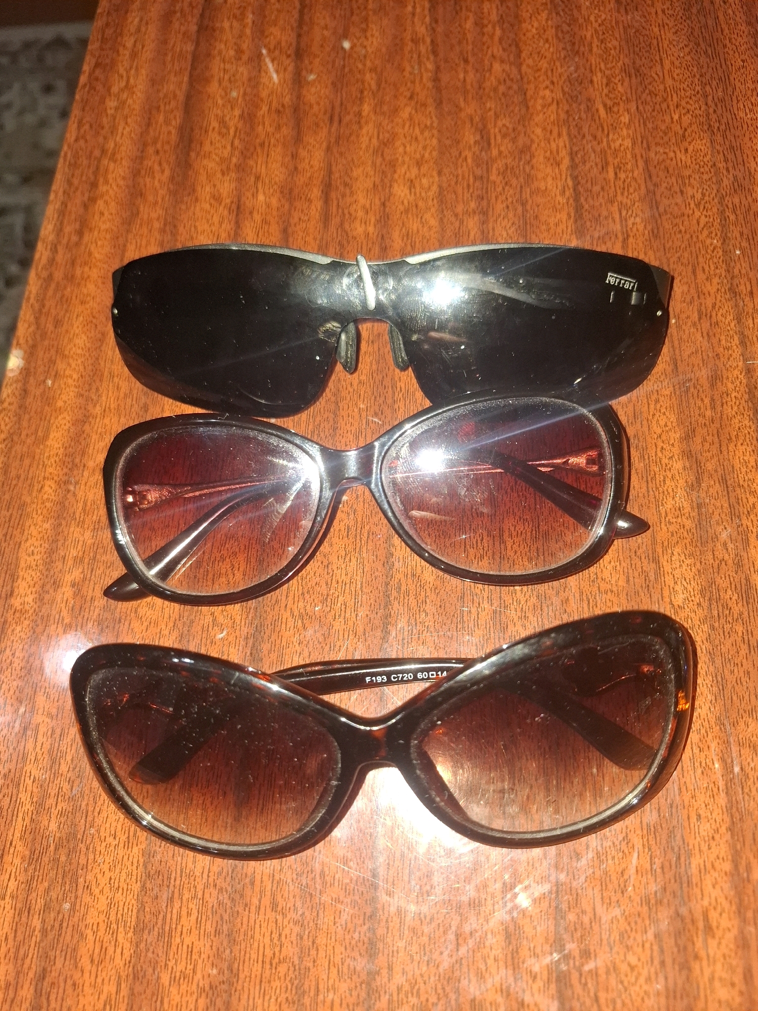 Three pairs of sunglasses are arranged vertically on a wooden surface. The top pair is black and appears to be a sporty style with a silver emblem on the right temple. The middle pair has black frames and reddish-brown lenses. The bottom pair has dark tortoiseshell-patterned frames and gradient brown lenses. The wooden surface has a visible grain. The scene is indoors, and the lighting suggests it may be artificial. No specific location in Almaty, Kazakhstan can be determined from the image.