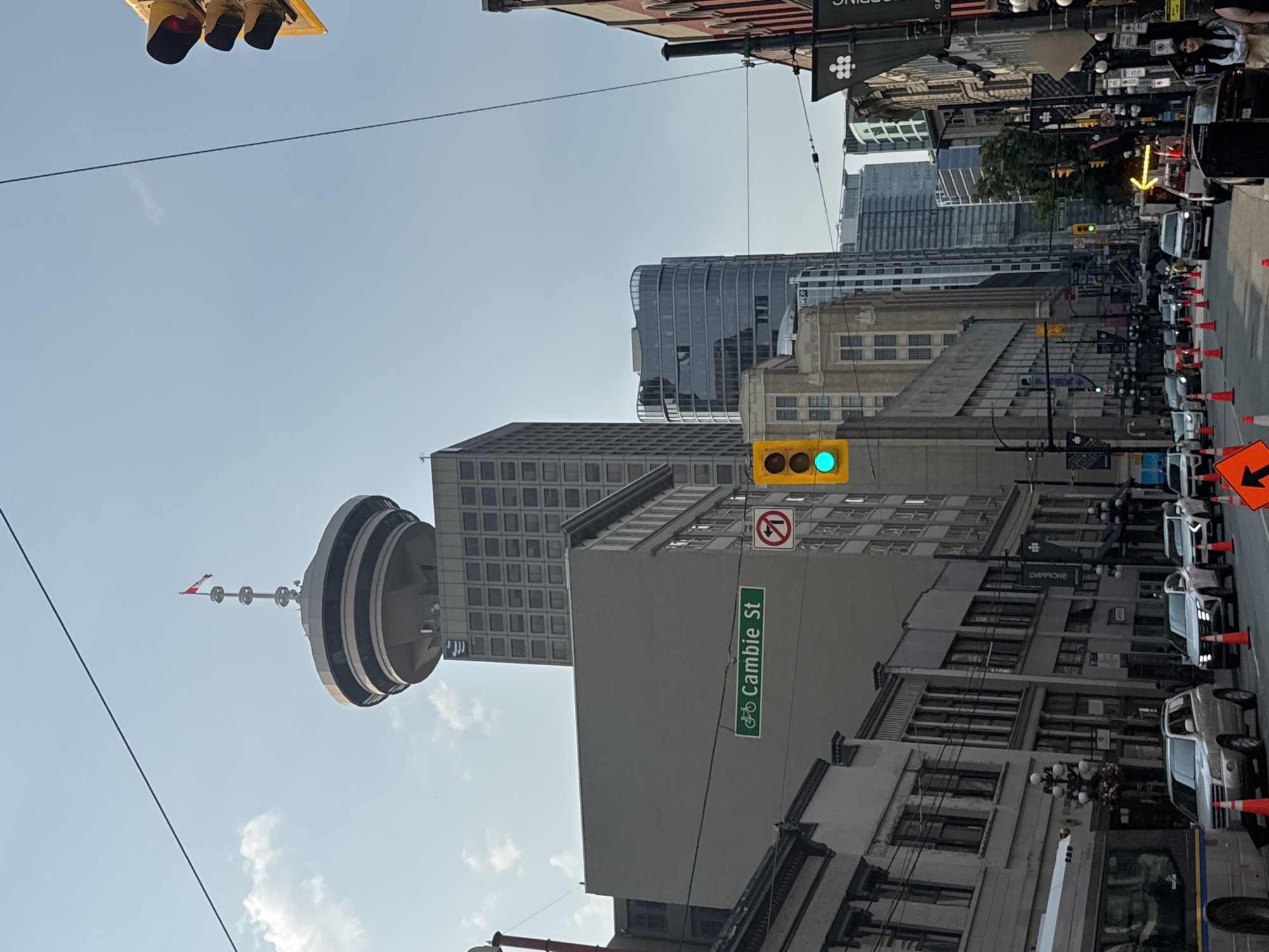 This is a daytime, eye-level, street-level shot of a downtown city scene, likely in Vancouver, Canada, given the presence of the iconic Vancouver Lookout tower, visible in the background. The scene is dominated by modern and older urban architecture, with tall buildings and a clear sky above.
In the foreground, a street is lined with vehicles, including a bus on the left and several cars and SUVs. Orange traffic cones are strategically placed on the road, suggesting ongoing construction or traffic management. A traffic light is visible, showing a green light. A street sign indicates "Cambie St" along with a bicycle symbol.
Several buildings are visible, showcasing a mix of architectural styles. On the left, older-style buildings with detailed facades line the street. Further back, a large, grey building stands prominently, followed by a modern, curved glass skyscraper. The Vancouver Lookout, a distinctive cylindrical observation tower, is perched atop a taller building in the background, its Canadian flag visible at the top.
People are present in the scene, though not in large numbers. A few individuals are walking or standing near the buildings and on the sidewalk. One person is visible inside the bus.
The overall impression is a bustling urban environment, with the green light and street signs indicating normal traffic flow despite the presence of traffic cones. The clear sky suggests fair weather.