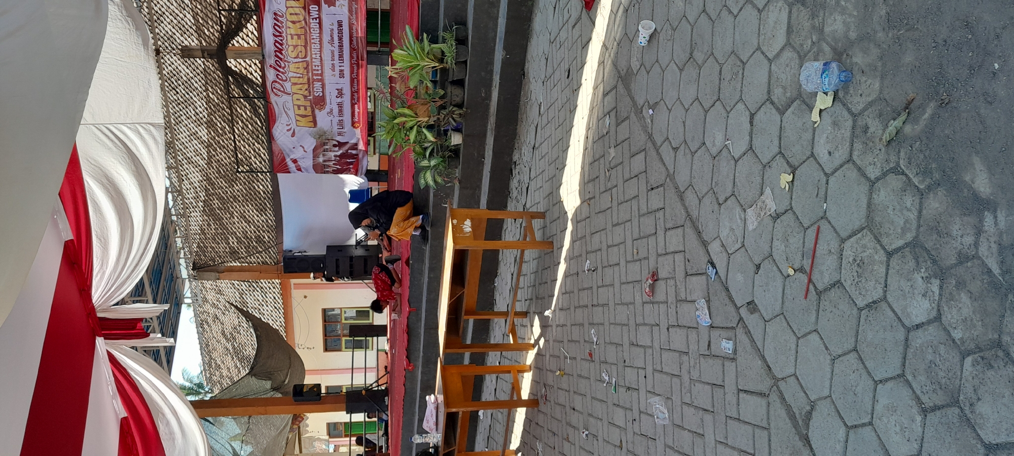 The image depicts an outdoor scene at SDN 1 Lemahbangdewo in Lemahbang Dewo, Indonesia. In the foreground, a paved area is littered with various pieces of trash including plastic bottles, wrappers, and straws. Two wooden desks with open compartments are placed on the paved area, with one further to the left and another to the right. In the midground, a red carpeted stage is visible, with steps leading up to it. On the stage, two individuals are present: a child wearing a red and black patterned shirt is kneeling and appears to be looking at something on the stage, while another person, dressed in a black headscarf and an orange-yellow outfit, is seated and looking down. Behind them, speakers are positioned on either side of the stage. A banner with text that appears to be in Indonesian is displayed on the stage's backdrop. The background shows a building with an orange-tiled roof and a white and pink facade, with windows visible. Overhead, a white and red fabric canopy is draped, supported by wooden poles.