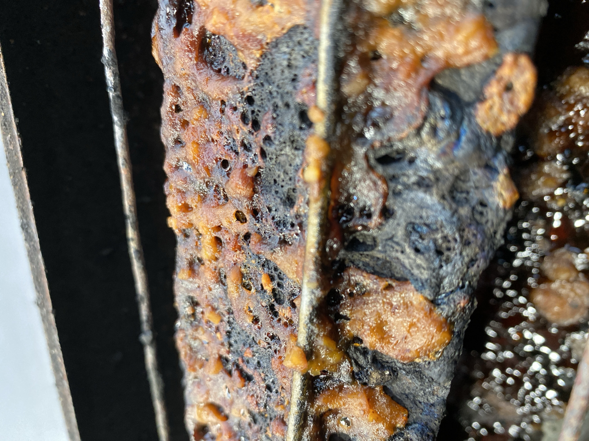 The image shows a close-up view of a greasy, charred metal grill rack, likely from a barbecue. The rack is covered in a thick layer of hardened, burnt residue, with some pieces of what appear to be remnants of food stuck to it. The metal itself is dark and blackened from the heat and grease. The background is out of focus but appears to be the inside of a grill or a similar metallic structure, with a lighter, out-of-focus surface above it. There are no people or other identifiable objects in the image. The scene is forensic in nature, focusing solely on the condition of the grill rack. The location in Kasihan, Indonesia, is not evident from the visual information provided in the image.