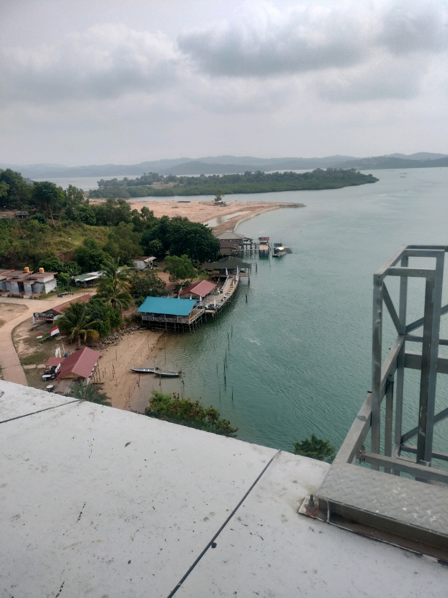 The image captures an aerial view of a coastal settlement in Batam, Indonesia, likely on a slightly overcast day. In the foreground, a grey concrete surface, possibly part of a building or platform, dominates the bottom of the frame, with some black cables and metallic structures indicating infrastructure.
The mid-ground reveals a cluster of buildings nestled along a sandy shore. Several structures are visible: a building with a red-tiled roof, another with a bright blue roof, and some smaller white buildings. A wooden pier extends into the turquoise water, with boats docked alongside. A small, light-colored boat is pulled up on the sandy beach. Palm trees and other lush greenery are scattered throughout the settlement, suggesting a tropical environment.
In the background, the calm sea stretches out towards a distant, tree-covered coastline with rolling hills. The water is a consistent greenish-blue, with subtle ripples indicating a gentle current. The sky is largely filled with soft, grey clouds. A metal railing structure is visible in the right portion of the frame, partially obscuring the view of the sea and the settlement below. The overall scene depicts a quiet, waterfront community, likely a fishing village or a small resort area, integrated with the natural coastal landscape of Batam.