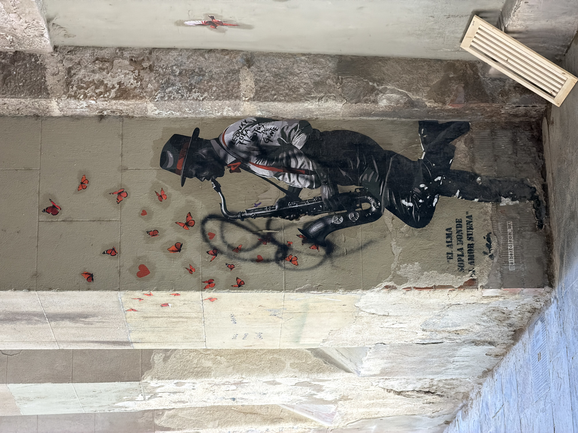 The image depicts a mural of a Black man playing a saxophone, rendered in black and white stencil art, on a textured, weathered wall in Barcelona, Spain. The mural is adorned with numerous small, red butterflies and some red hearts scattered across the wall. The saxophonist wears a fedora hat, a red bowtie, suspenders, and a shirt with handwritten text on it. Below the saxophonist, a Spanish phrase is stenciled: "EL ALMA SOPLA DONDE EL AMOR SUENA" which translates to "The soul blows where love sounds." The artist's tag, "@albertonleon_art," is visible at the bottom right of the mural.
The mural is situated within an arched passageway, with rough, light-colored stone pillars on the left and a rougher, darker stone corner on the right. The wall itself is a light beige color, showing signs of age and damage, with visible patches of brick. There is also some graffiti on the wall to the left of the mural. On the floor, there is a section of light-colored pavement and a few drains. To the right of the mural, leaning against the wall, is a simple wooden slatted object. The overall environment suggests an outdoor urban setting, possibly a public walkway or courtyard.