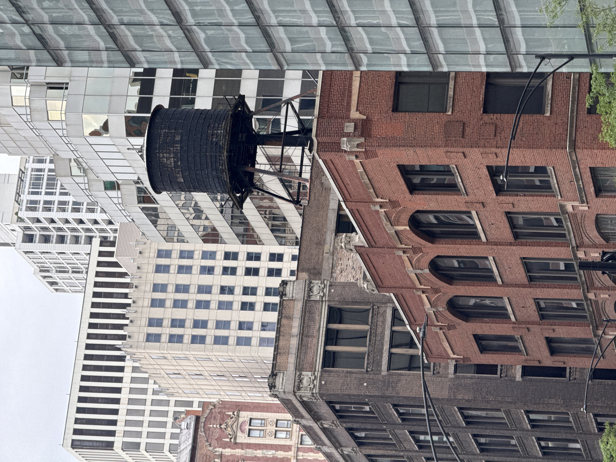 An urban cityscape showcases a dense collection of buildings of varying architectural styles and heights, set against a bright, overcast sky. In the foreground, a red brick building with arched windows and decorative stone elements occupies the lower portion of the frame. To its left, a darker brick building exhibits a more industrial aesthetic with numerous rectangular windows.
Dominating the mid-ground is a large, dark, cylindrical water tower perched atop a metal support structure. This water tower is positioned in front of a lighter-colored building with a grid-like pattern of windows, which is itself behind a modern glass-fronted skyscraper. Further in the background, several other multi-story buildings are visible, including one with vertical dark and white stripes and another with a checkered facade. The scene suggests a bustling metropolitan environment, likely in Chicago, United States of America, given the characteristic architectural elements and the presence of water towers. There are no visible people in the image. The overall impression is one of urban density and architectural layering.