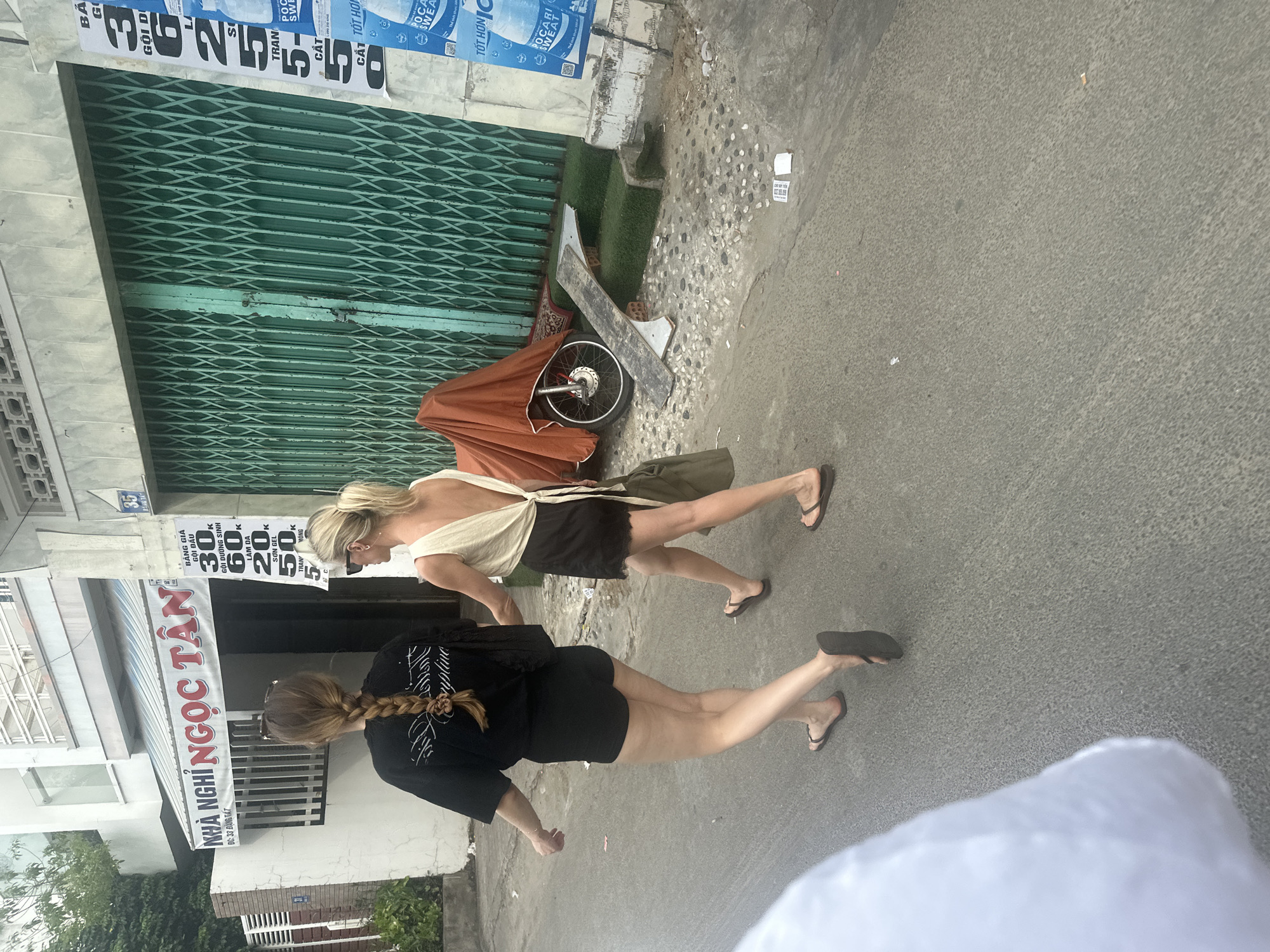 Two women walk along a paved street in Bac Nha Trang Ward, Vietnam. One woman has blonde hair in a bun and is wearing a cream-colored top and a black skirt. The other has long brown hair in a braid, wearing a black t-shirt and black shorts. They are both wearing flip-flops. A closed green metal accordion-style gate is to the left. Next to the gate is a dilapidated moped covered with an orange tarp. A sign for "Nhà Nghỉ Ngọc Tân" can be seen on a building to the left, along with a price list for services. Several advertisements in Vietnamese are posted along the wall above the metal gate.