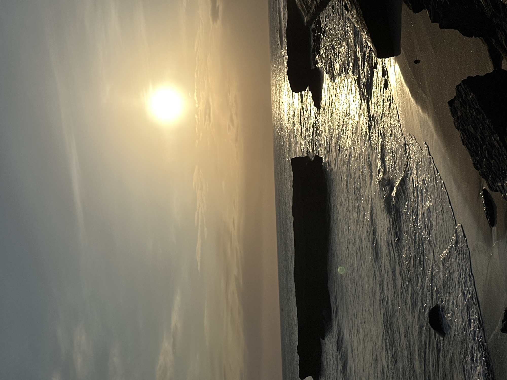 A coastal view features a bright sun visible in a hazy sky. To the right, the ocean surface reflects light, and a rocky coastline is visible along the water's edge. The image appears to be taken at or near sunset, with warm, muted tones dominating the composition. The location is North Kuta, Indonesia.