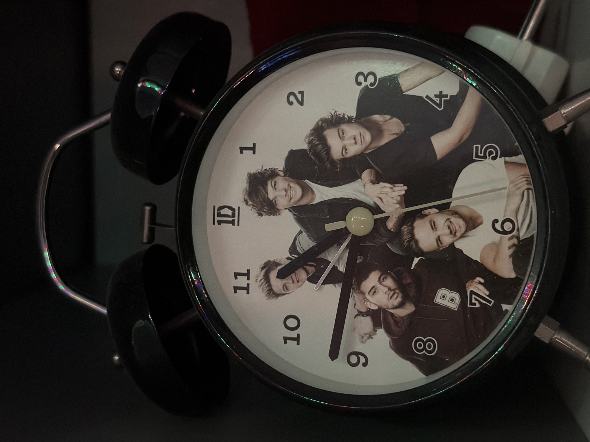 The image shows an alarm clock with a black frame and bells, the clock face is white with black numerals 1-12, and the background is a photograph of the band One Direction. The time displayed is approximately 8:51. The clock is indoors, sitting on a dark surface. The location, Varna, Bulgaria, cannot be confirmed from the visual information provided.