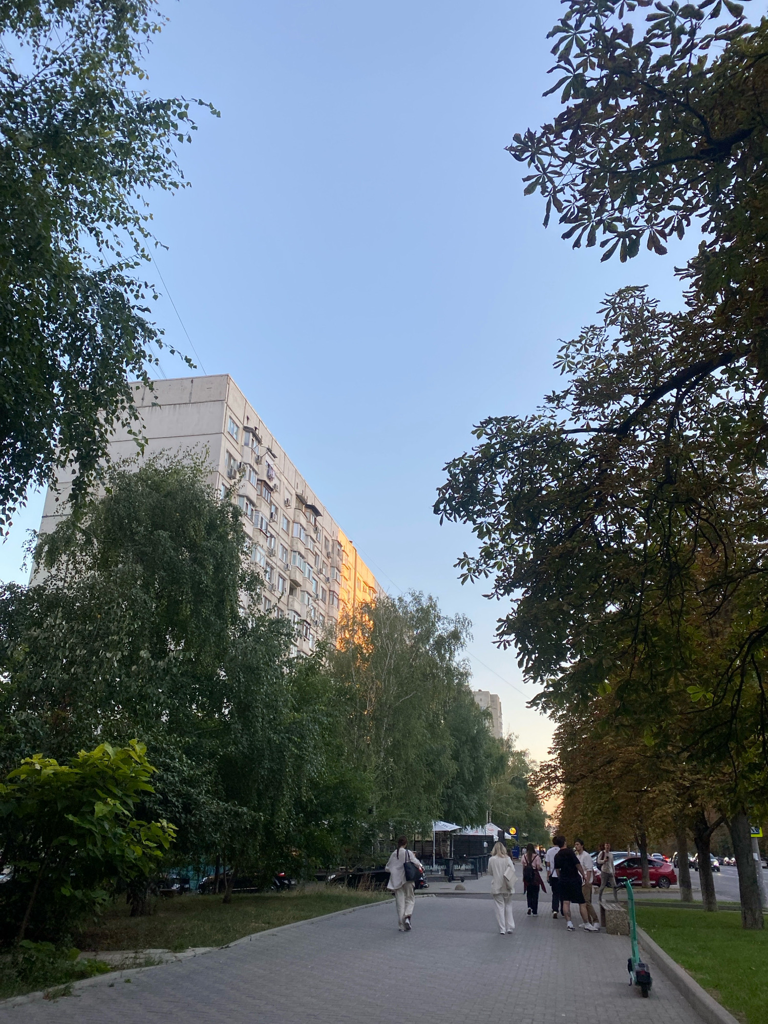 A group of people walk along a paved sidewalk next to a street lined with trees and a multi-story apartment building in Chișinău, Moldova. The apartment building is tall and light-colored with many windows and balconies. The trees have green leaves, and some are beginning to show signs of autumn colors. The sky is clear and light blue. The sidewalk is wide and appears to be made of concrete pavers. Several cars are visible on the street. One person is on an electric scooter. The scene appears to be taken in the late afternoon or early evening, as the sun is casting long shadows and highlighting the side of the building.