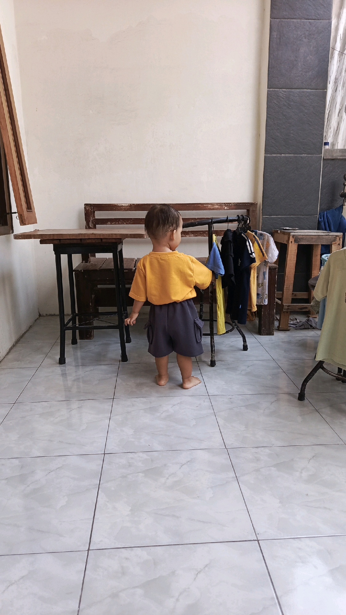 In Pekauman Kulon, Indonesia, a young child stands barefoot on a tiled floor, facing away from the camera. The child is wearing a bright orange t-shirt and dark gray shorts with cargo pockets. To the child's right is a clothing rack with several garments hanging on it. Behind the child, a wooden bench is visible against a textured white wall. To the left of the bench is a wooden table with black metal legs. The environment appears to be an outdoor or semi-outdoor space, possibly a patio or veranda, as indicated by the tiled floor and the presence of laundry drying on a rack.