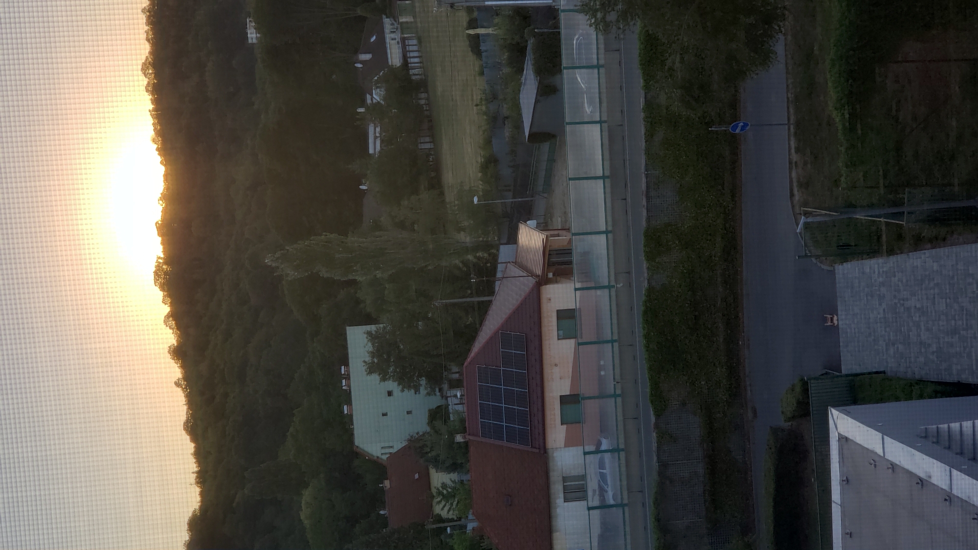 The image shows a residential area during sunset. In the foreground, there is a roof of a modern building. Beyond that, a street with a few trees and a blue traffic sign. Further in the background, a hill covered with dense trees, with a few houses scattered amongst them. A house with solar panels is visible on the slope of the hill. The sky is orange and yellow, indicating the sun is setting. The scene is viewed through a window with a screen.