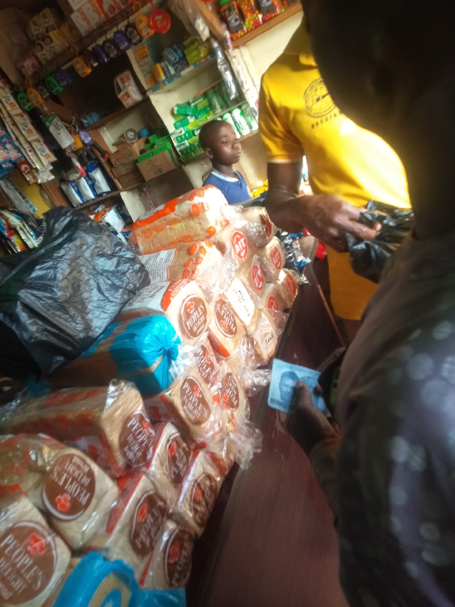 In a bustling corner store in Ejule, Nigeria, a transaction is underway. The counter is stacked with loaves of "People's Delight Bread," neatly packaged. A person in a polka-dot shirt is paying with a blue banknote. A vendor in a yellow shirt handles a black plastic bag. A boy watches in the background amidst shelves stocked with various goods.