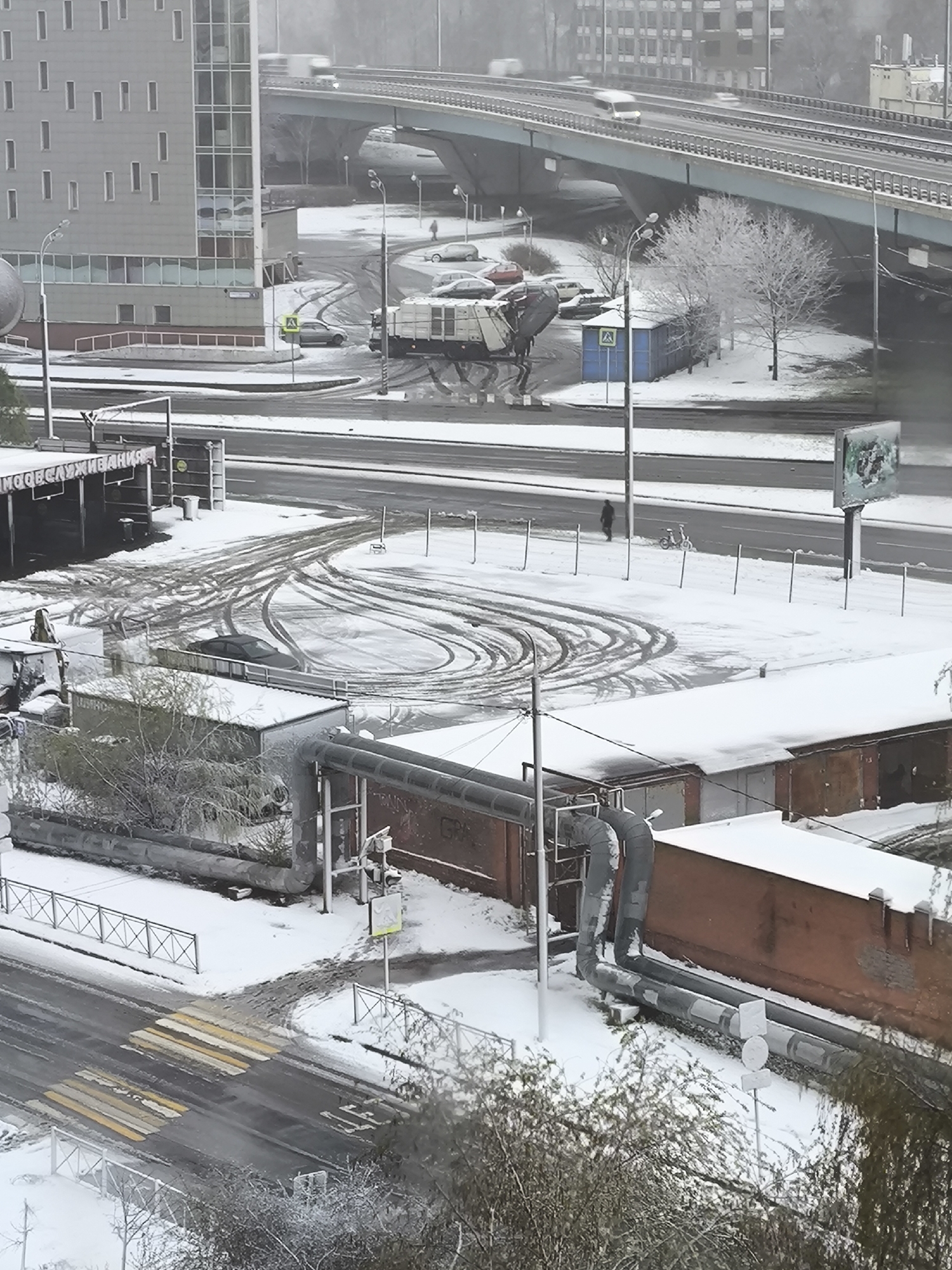 This image captures an urban landscape in Khimki, Russia, on an overcast winter day. The entire scene is covered in a fresh layer of snow, dusting roadsides, buildings, and trees.
In the foreground, large industrial pipes, partially covered in snow, run alongside brick and concrete buildings. A zebra crossing with faded yellow and white stripes is visible on a lower road. An open, snow-covered area features distinct, looping tire tracks, indicating recent vehicle activity. To the left, a modern, tall grey building with numerous small, irregularly placed windows rises above the street level.
The middle ground is dominated by an elevated multi-lane highway or bridge, across which blurred vehicles are seen moving, suggesting motion. Beneath the bridge, a large, distinctive multi-axle vehicle, possibly a specialized industrial truck or machine, is prominently parked, leaving deep tracks in the snow. Adjacent to it are a blue shed-like structure and several parked, snow-covered cars. A standard pedestrian crossing sign is also visible in this area.
A lone figure walks on the snow-dusted sidewalk near the main road in the mid-right section of the image, while another faint figure can be seen under the bridge. A small, white child's bicycle leans against a fence near the road. The bare trees throughout the scene are heavily frosted with snow, contributing to the cold, wintery atmosphere.
Visible text includes "ООО ОБСЛУЖИВАНИЯ" (LLC SERVICE) on a sign near a building entrance in the lower-left portion of the image. Partial Cyrillic letters like "ГРА" (GRA) are discernable on a brick wall, likely graffiti. The content of a large billboard near the road is unreadable.