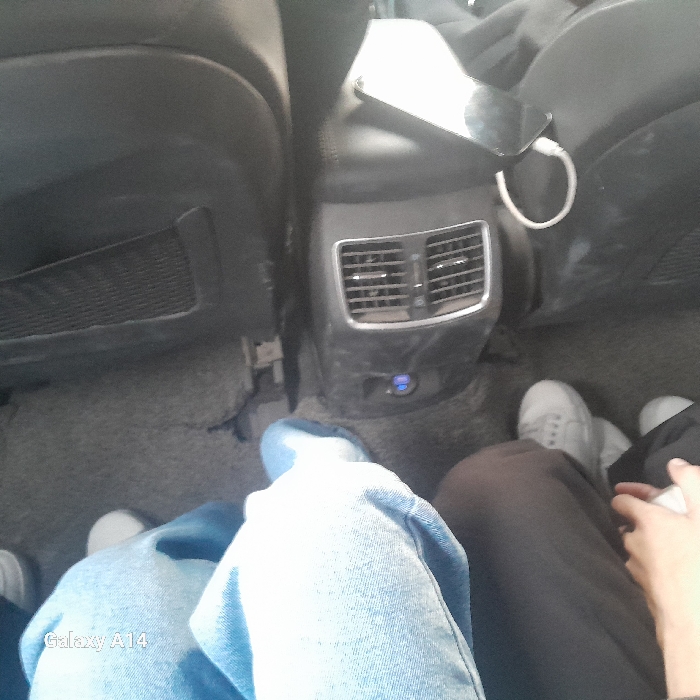 The image shows the interior of a car, specifically the back seat area. Two car seats are visible, with a center console between them that has vents and a phone charging on top. Two people are sitting in the back, one wearing jeans and blue sneakers, the other wearing dark pants and gray sneakers. The overall lighting is neutral, and the image appears to be taken from a slightly elevated angle.
