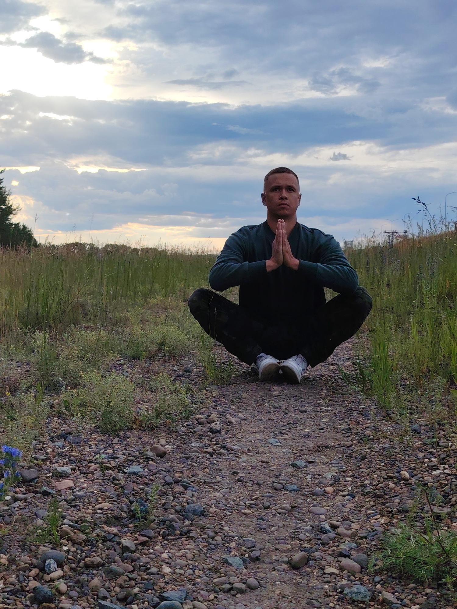 A man with short hair and a serious expression is meditating in a squatting position on a gravel path. He is wearing a dark long-sleeved shirt and dark pants, and white sneakers. His hands are pressed together in front of his chest. He is surrounded by tall green grass and wildflowers. In the background, there is a cloudy sky with the sun peeking through. The image appears to have been taken outdoors in a natural setting, possibly in the city of Chaikovsky, Russia, given the vegetation and general landscape.