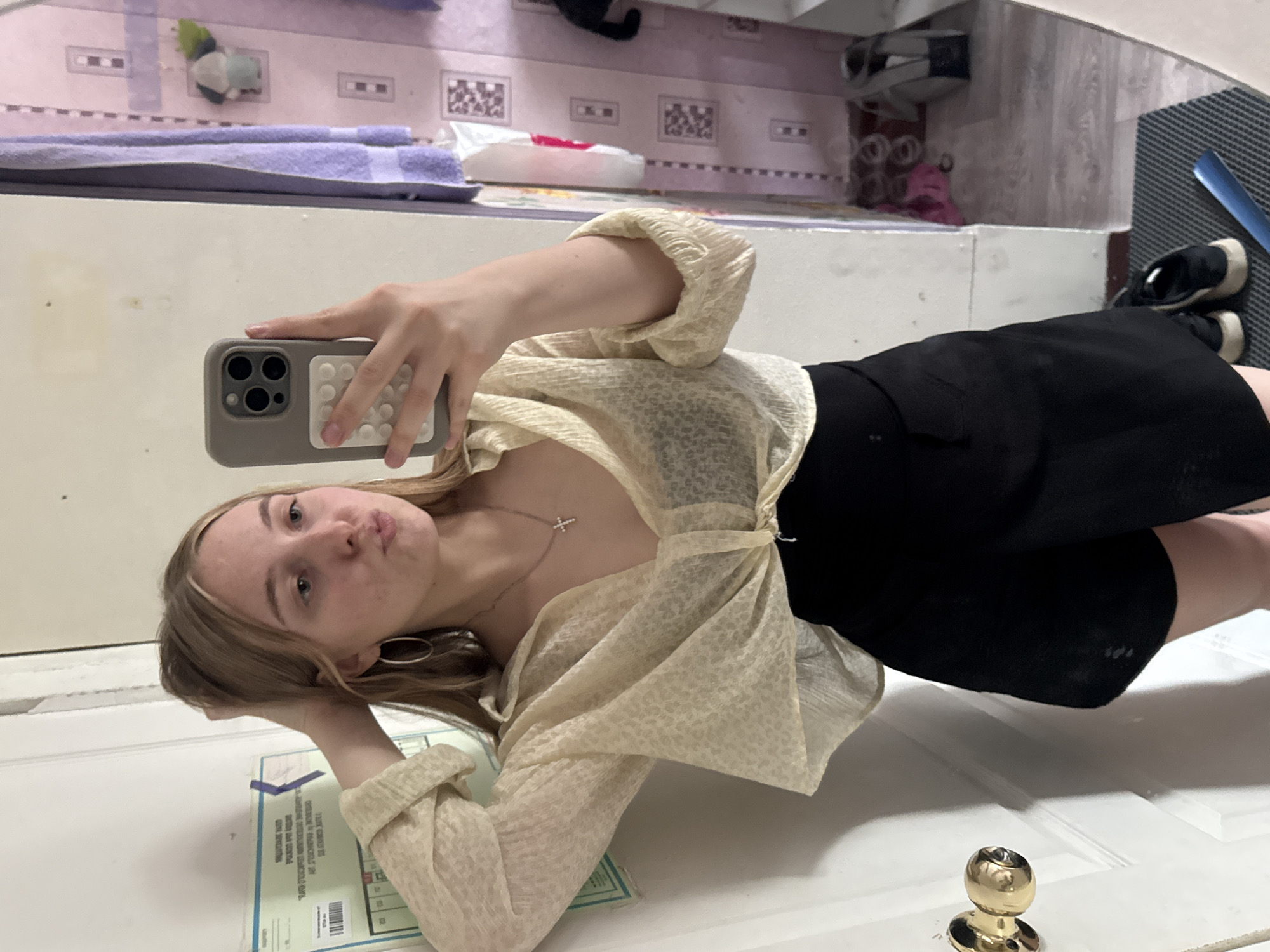 The image is a selfie taken by a young woman indoors, likely in a residential setting. She has blonde hair and is wearing a light-colored, slightly sheer blouse tied at the waist and a black skirt. A silver cross necklace is visible. She is holding her phone up to take the picture, and her other hand is resting on her head. In the background, there is a door with a gold doorknob, a mirror reflecting a cluttered space with what appears to be towels and possibly shelving, and a portion of a wall with some papers attached. The scene appears to be a casual, personal moment captured by the subject. There is no information in the image to suggest a specific location in the city of Perm, Russia.