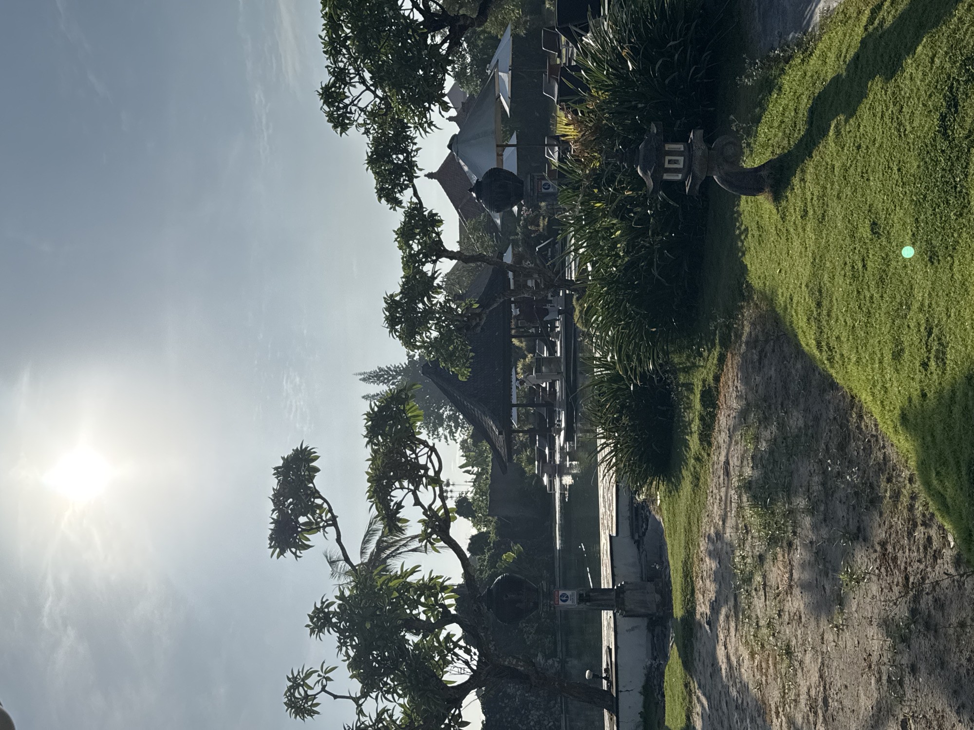 The image shows a sunlit outdoor scene in Jimbaran, Indonesia. The foreground features sandy ground transitioning to green grass on the right side. Several trees with green leaves are visible, some framing a view of a building with a multi-tiered roof and open structure behind. Elements suggesting a resort or garden setting are present, including landscaped greenery and potentially outdoor seating. The sky is bright with the sun visible in the upper left corner. No people are seen in the image.