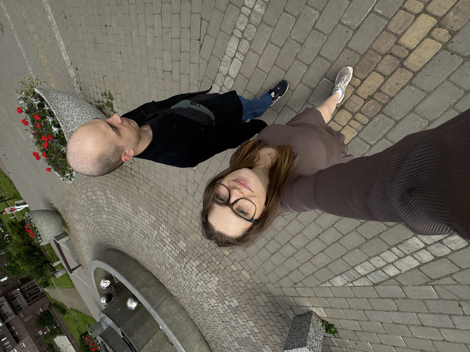 The image shows two people, a man and a woman, standing on a paved walkway. The man, on the left, has a shaved head and is wearing a black jacket over a dark t-shirt and blue jeans. He has a black fanny pack around his waist. The woman, on the right, is wearing glasses and a brown dress. She is holding the camera to take a selfie.
They appear to be walking on a public street or plaza, with patterned paving stones underfoot. To the left of the man, there is a large planter filled with red and white flowers. In the background, there is a curved concrete structure with decorative elements and some greenery. Further in the distance, buildings and trees are visible.
The lighting suggests it is daytime, likely overcast, as there are no strong shadows. The overall mood seems casual and relaxed, as they are taking a selfie together. The presence of cobblestone-like paving, planter boxes, and the architectural style of the background elements are consistent with urban settings in Saint Petersburg, Russia. No visible text is present in the image.