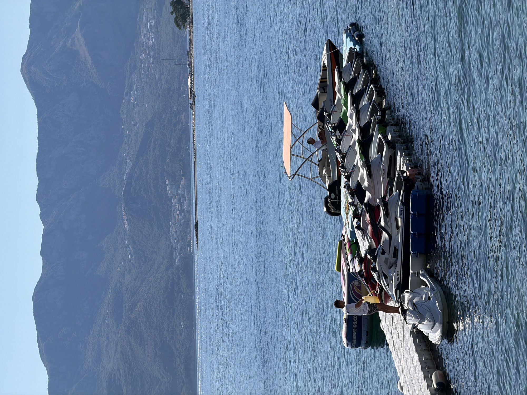 This image depicts a sunny day at a coastal location, likely the city of Budva, Montenegro, given the mountainous backdrop often seen in the region. The scene features a wooden dock extending into calm, blue water. Moored to the dock are numerous jet skis lined up in rows, suggesting a rental or activity center. One white jet ski is partially submerged in the water at the front of the dock. Behind the jet skis, a small boat with an orange canopy is also docked. A man in a white t-shirt and patterned shorts stands on the dock, holding a yellow container, possibly refilling one of the jet skis. Another person is seated in the small boat. In the background, a hazy mountain range covered in green vegetation rises above the water, with scattered buildings visible on its slopes. The sky is clear and bright blue.