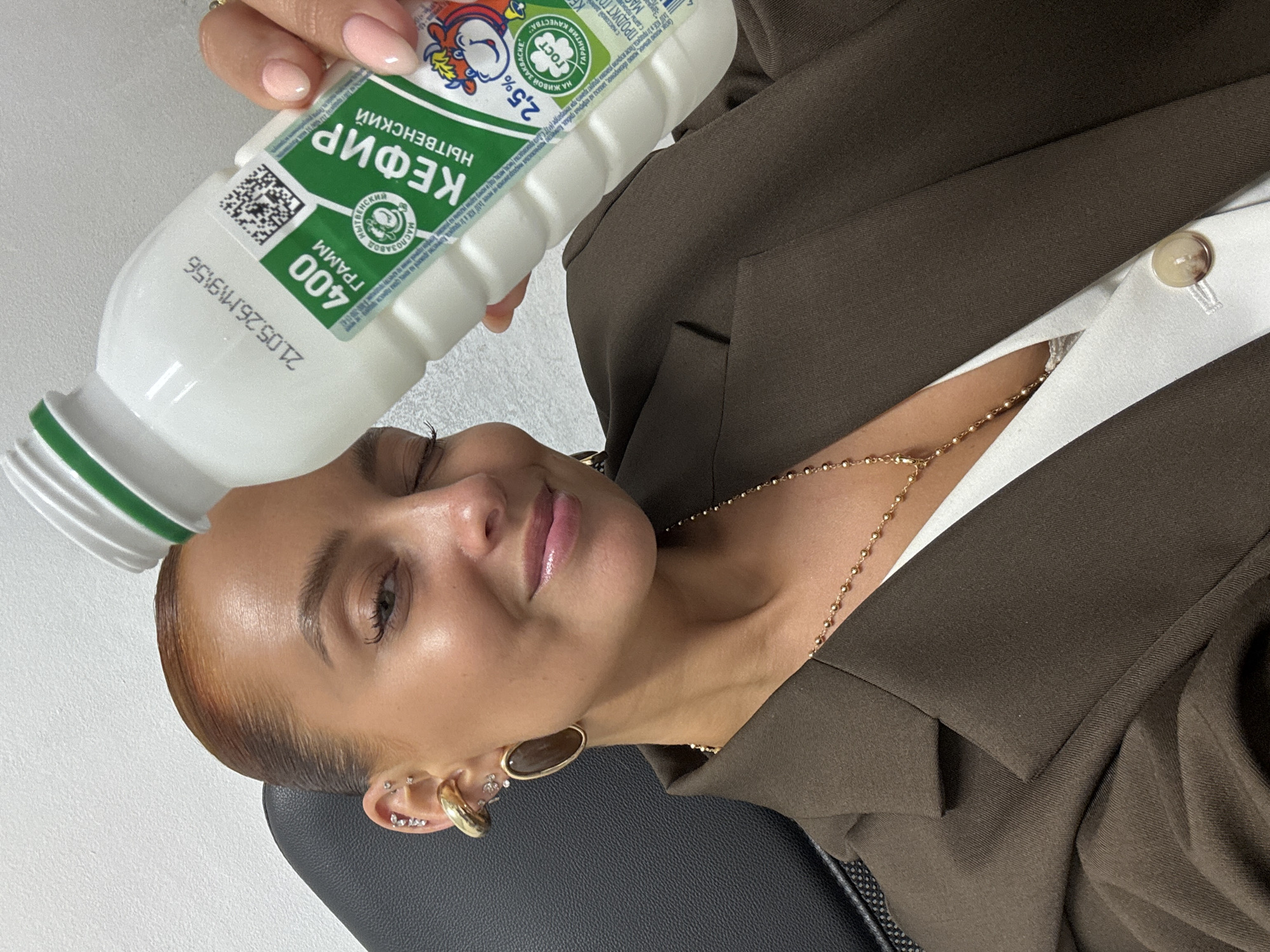 A woman is smiling and holding a bottle of kefir on her forehead. The label on the bottle is in Russian and indicates the product is Kefir Nytvenskiy 2.5% fat, weighs 400 grams, and has a production date of 21.05.26. The location is the city of Chaikovsky, Russia. The woman is wearing a brown blazer and a white top with a gold necklace. She also has several piercings in her ear, including a large gold hoop. She is seated in a black chair.