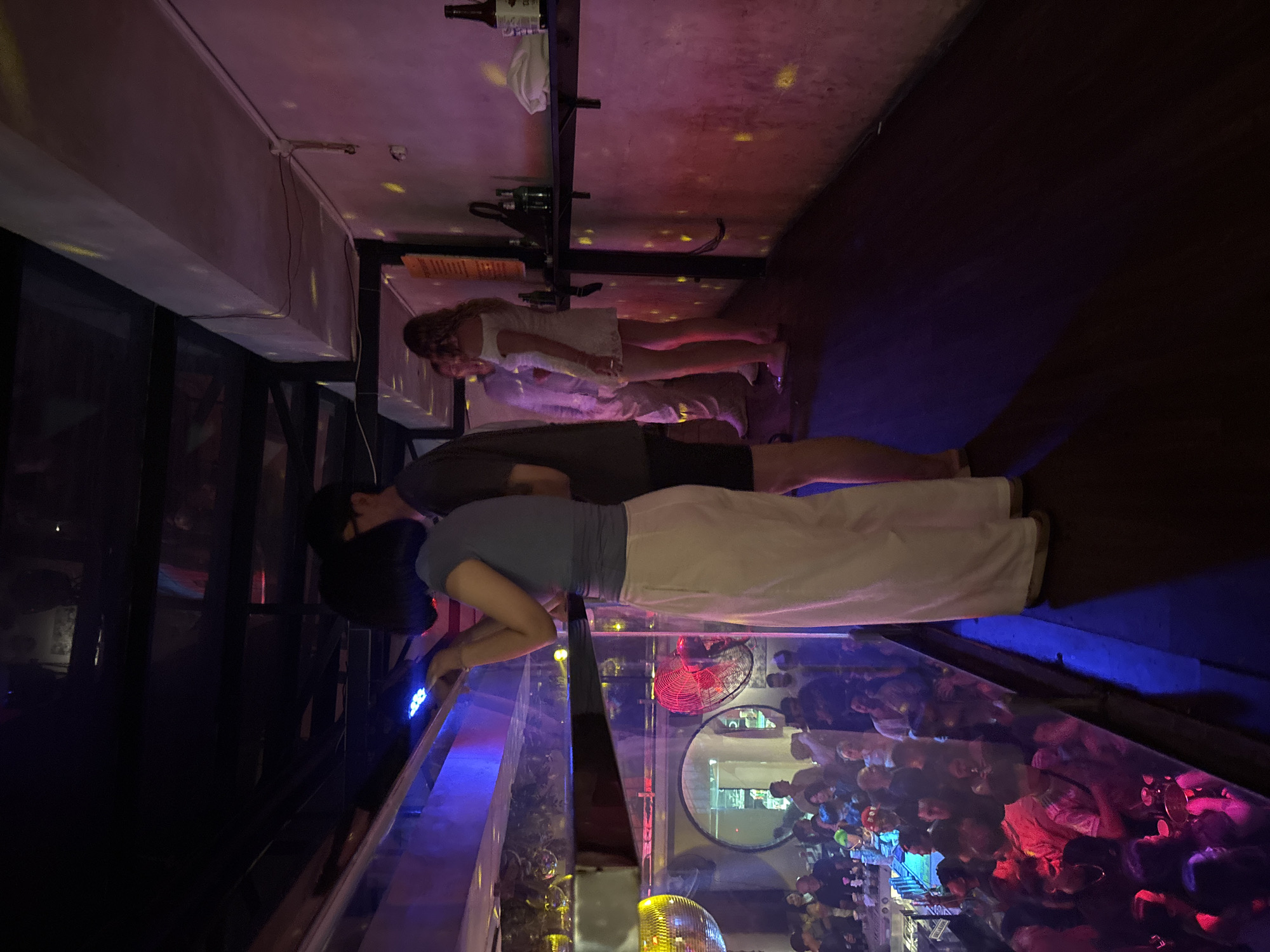 In a dimly lit, multi-level establishment in Da Nang, Vietnam, likely a nightclub or bar, a group of people are gathered. In the foreground, a woman wearing a blue t-shirt and white pants stands facing a glass barrier, her hand raised towards a strip of blue LED lights. Beside her, a man in a dark t-shirt and shorts is also looking towards the barrier. Below them, through the glass, a crowded scene unfolds with many people seated at tables, illuminated by colorful lights. Disco balls are visible, and a red electric fan is mounted on the wall. Further back, on a raised level, another group of people are standing, including a woman in a white dress. The walls are a textured pink and purple, with scattered yellow lights adding to the ambient lighting. Shelves are present, holding bottles and a white cloth. The flooring is dark.