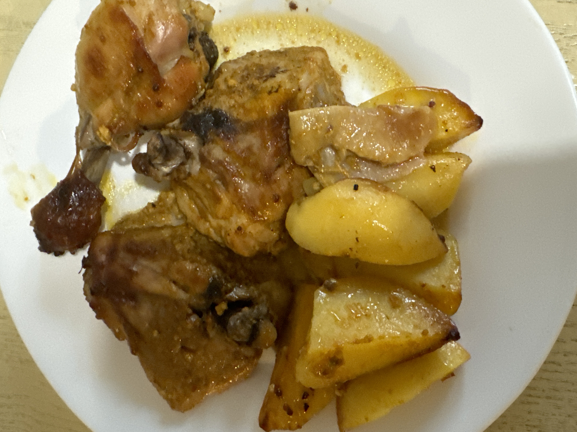 The image shows a white plate with roasted chicken pieces and roasted potatoes. The chicken appears to be a mix of light and dark meat, with some areas slightly charred. The potatoes are cut into wedges and have a golden-brown color, suggesting they are well-cooked. There is some liquid or sauce on the plate, possibly pan drippings from the cooking process. The overall setting appears to be a home or restaurant setting. No information can be derived from the image to identify its location in the city of Chaikovsky, Russia.