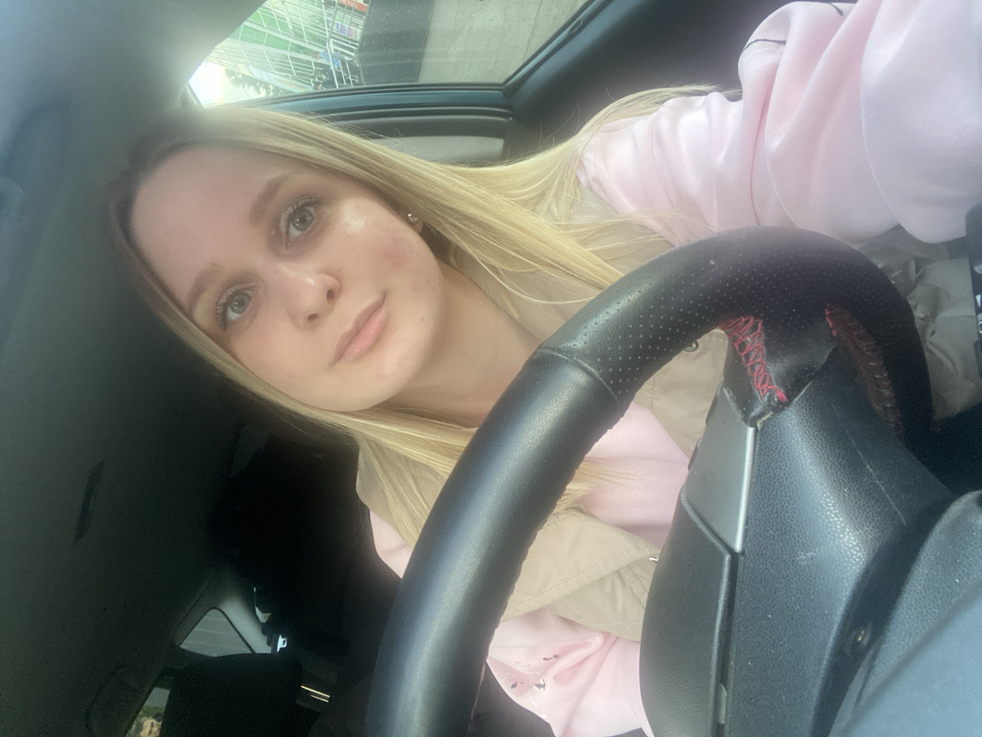 A young woman with blonde hair is sitting in a car, behind the steering wheel. She is wearing a pink sweatshirt and a light beige jacket. The steering wheel is black with some red stitching visible. Through the open sunroof, what appears to be a shopping center structure is partially visible. Based on the architectural style of the visible part of the building and the presence of shopping trolleys, the location is likely a shopping center in Chaikovsky, Russia.
