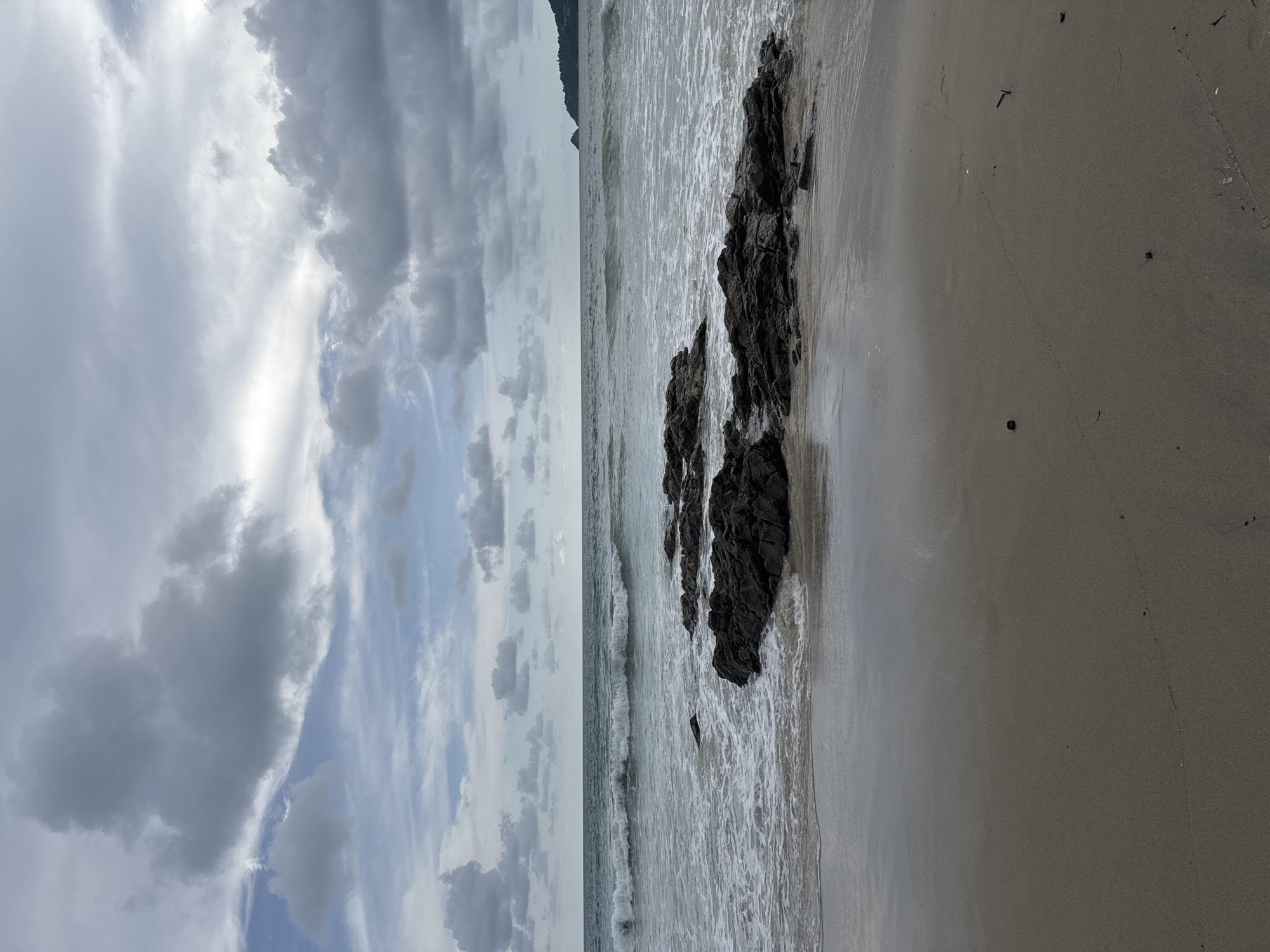 The image depicts a serene beach scene in Patong, Thailand, under a dramatic, cloudy sky. Dark, rugged rocks are partially submerged in the incoming tide, creating a natural barrier against the gentle waves. The water is a deep blue-grey, reflecting the overcast conditions. In the distance, a faint outline of a hill or headland is visible on the horizon, characteristic of the Patong coastline. The sandy shore in the foreground is damp and reflects the light, with a few scattered pieces of debris. There are no people or animals visible in the frame. The overall atmosphere is one of tranquility and natural beauty, hinting at the approaching or receding end of a storm.