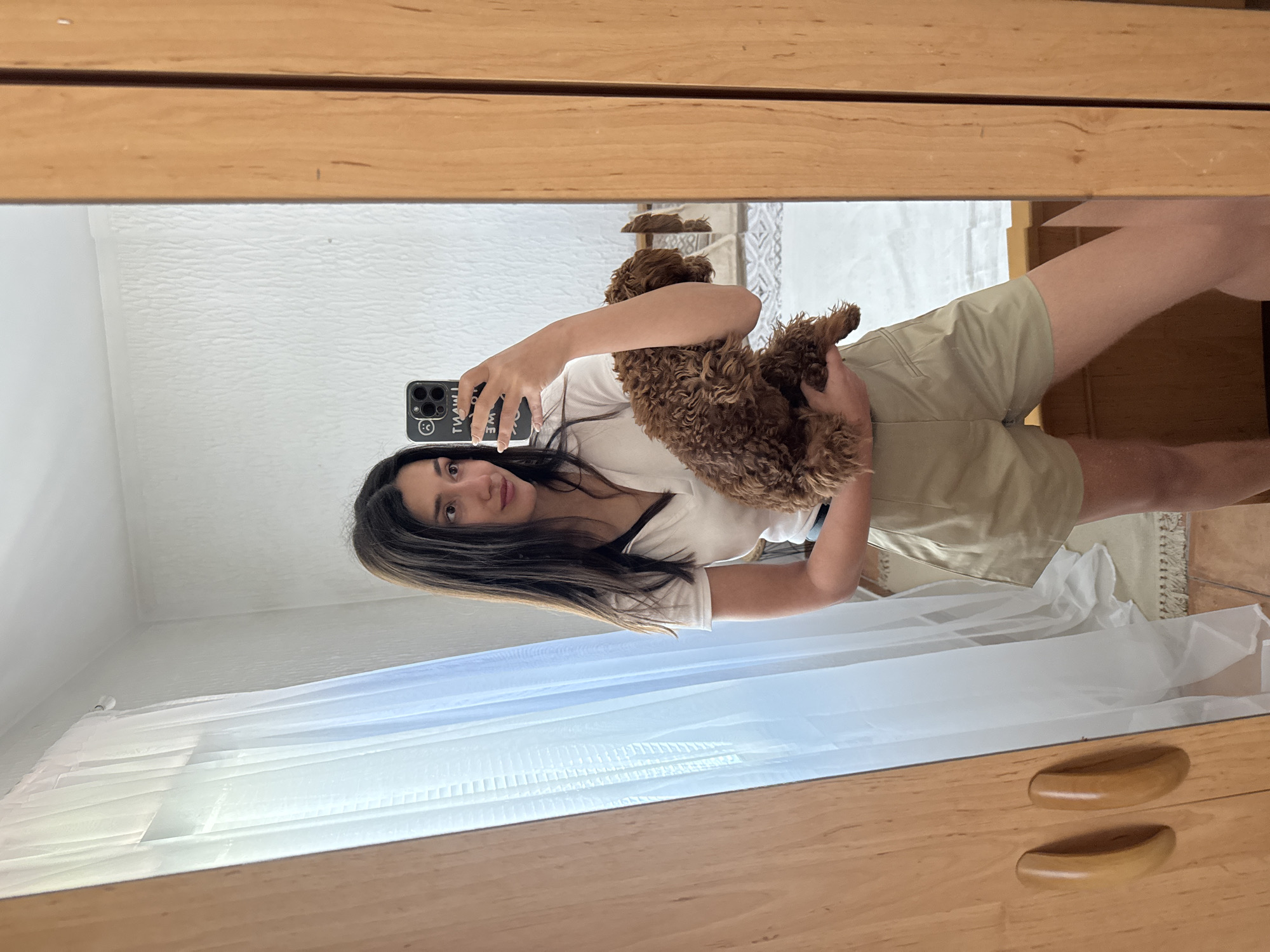 A young woman is depicted taking a mirror selfie, holding a small, fluffy brown dog in her arms. She has long dark hair and is dressed in a white short-sleeved top and light beige shorts. Her expression is neutral as she looks towards her smartphone, which she holds with her right hand. The phone is black with a white case, featuring black text that reads "I WANT YOU" (reversed in the mirror) along with several emojis, including a sad face and an X-eyes smiley face.
The small dog, a brown curly-haired breed resembling a poodle or doodle, is nestled comfortably in her left arm.
The reflection in the mirror shows a bright indoor setting, likely a bedroom or living space, with white textured walls. A large window covered with sheer white curtains is visible in the background, indicating ample natural light, suggesting it is daytime. The mirror itself is framed by light-colored wooden cabinetry, possibly a wardrobe or dresser, which also features two curved wooden handles in the lower right portion of the reflection. A light-colored rug or carpet is visible on the floor in the reflected background.
The scene portrays a casual, relaxed moment at home in Sutomore, Montenegro.