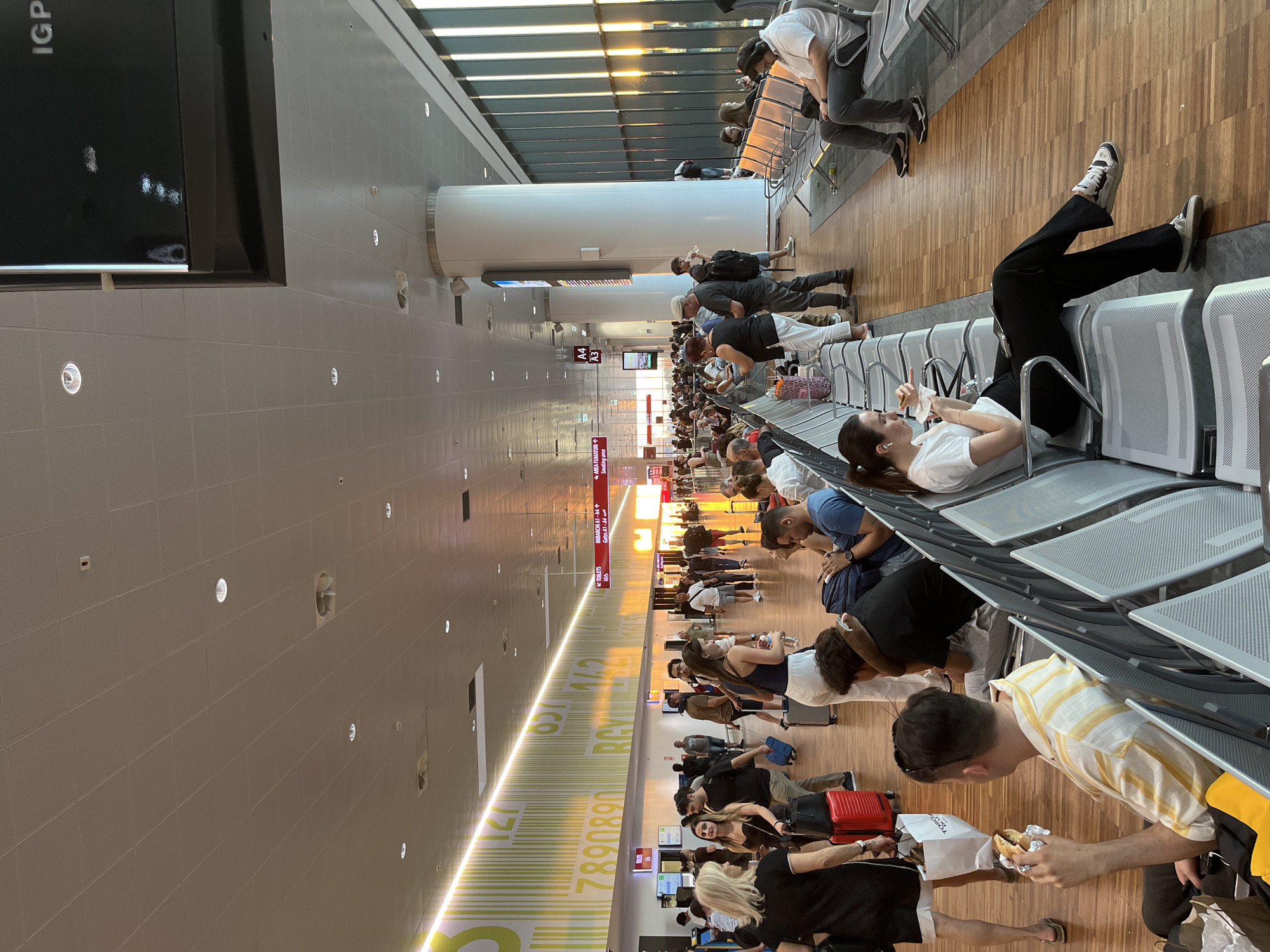 The image depicts an interior scene within Orio al Serio International Airport in Orio al Serio, Italy. The environment is a spacious, modern airport terminal with a high ceiling, featuring tiled flooring and large windows along one side. A prominent element of the architecture is the presence of large white cylindrical pillars. The lighting suggests it is late afternoon or early evening, with sunlight streaming through the windows.
A large number of people are present, many of whom are seated on rows of airport benches, engaged in various activities such as waiting, checking phones, eating, or talking. Others are standing, walking, or pulling luggage. The individuals are diverse in age and gender, dressed in casual attire.
Key objects visible include luggage, suitcases, backpacks, shopping bags, and personal electronic devices. Overhead, there are lights, ventilation systems, and signage. Notably, large decorative graphics, resembling bar codes and numbers, are present on the wall in the background. Directional signs indicate "TOILETS" and gates. The overall atmosphere appears to be one of a busy transit hub.