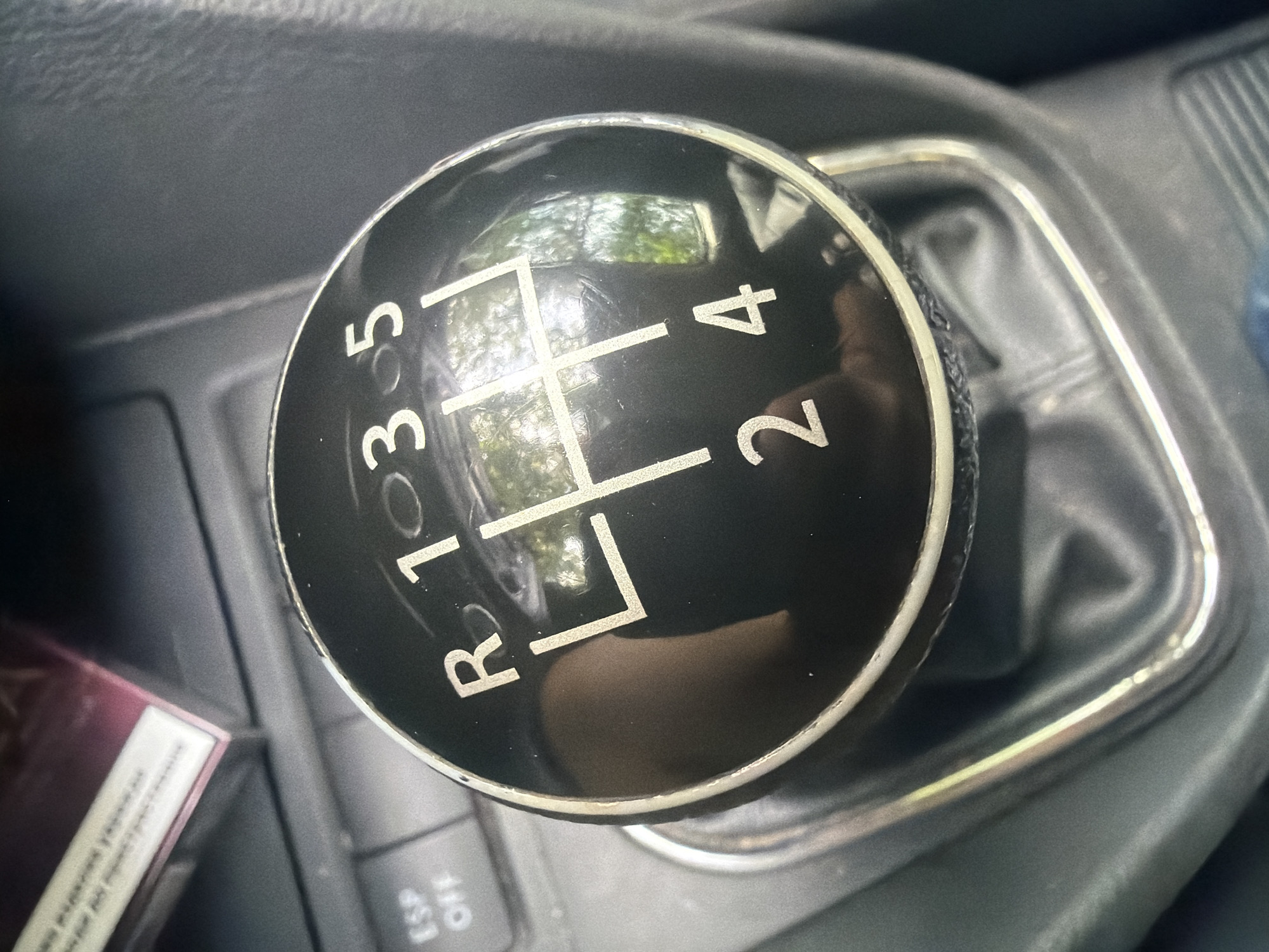 The media file captures a close-up, high-angle view of the center console of a car, specifically focusing on a manual gear stick, located in Iskar, Bulgaria. The primary subject is a black, round gear shifter knob with a silver trim, indicating a 5-speed manual transmission with reverse (R, 1, 2, 3, 4, 5) clearly marked in white lettering. The gear pattern is etched into the top of the knob.
The gear stick is surrounded by a wrinkled black leather or synthetic gear boot (gaiter), which is enclosed by a rectangular silver-colored bezel. The surrounding car interior is made of dark grey or black textured plastic, showing some signs of use.
Reflected on the glossy surface of the gear knob and the car interior elements are what appear to be trees and bright daylight, suggesting the image was taken during the daytime. To the left of the gear stick, a partially visible box, possibly a pack of cigarettes, with white and purple/red packaging, is tucked into a storage compartment. Below the gear stick, a button labeled "ESP OFF" is partially visible on the console. There is no direct activity depicted, but the context is that of a vehicle's interior. No people are visible.