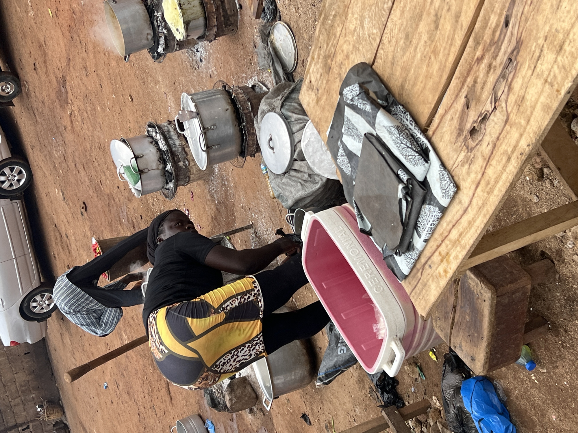The image shows a woman cooking outdoors in Gura Top, Nigeria. She is squatting next to several large pots sitting on top of open fires. She is wearing a black shirt and patterned yellow shorts. Nearby is a wooden table with a patterned black and grey fabric on it, as well as a pink plastic tub. A white car is visible in the background to the left. The ground is dirt and there are various other objects scattered around the scene.