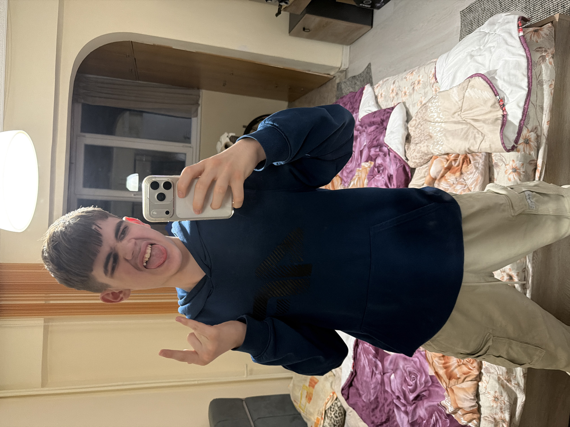 An indoor, mirror selfie captures a young male making a playful and mischievous face, sticking his tongue out. He is seen horizontally in the frame, holding a silver-colored iPhone with a clear case in his right hand to take the picture. His left hand is raised, forming a "rock on" gesture with his index finger and pinky extended.
The subject wears a dark blue hooded sweatshirt with a large, stylized black logo on the chest, and light beige cargo pants. He has short, dark hair with a fringe.
The background shows a domestic interior, likely a bedroom or living area. To the left, a bright white lamp shade is visible. Behind the person, a bed is covered with an assortment of textured and patterned blankets and comforters, featuring floral designs in shades of purple, beige, and white. An arched doorway in the background reveals a window with blinds. A dark wooden shelf or cabinet is mounted on the wall. The floor appears light-colored, with a patterned rug nearby. The lighting suggests an indoor setting, possibly with a mix of natural and artificial light. No legible text is visible other than the phone's camera array and the indistinct logo on the hoodie.