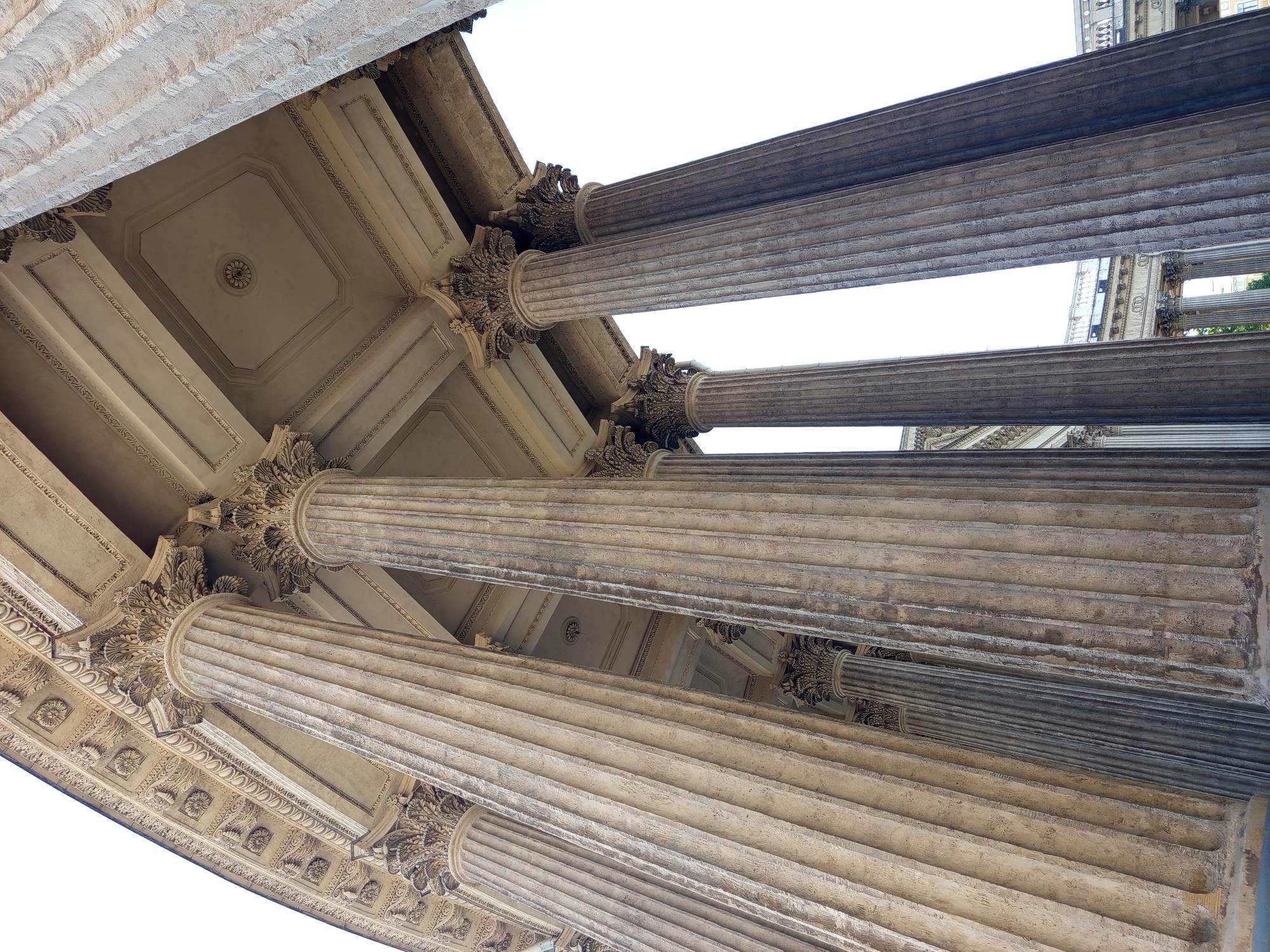 This is a low-angle, upward shot of the Kazan Cathedral in Saint Petersburg, Russia. The image focuses on the cathedral's colossal Corinthian columns, which are fluted and made of stone. The columns are arranged in a curved colonnade, with the perspective emphasizing their height and grandeur. In the background, glimpses of the building's ornate facade and ceiling are visible, featuring intricate decorative carvings and geometric patterns. The sky is bright and overcast, providing even lighting. There are no people or discernible actions in the frame.