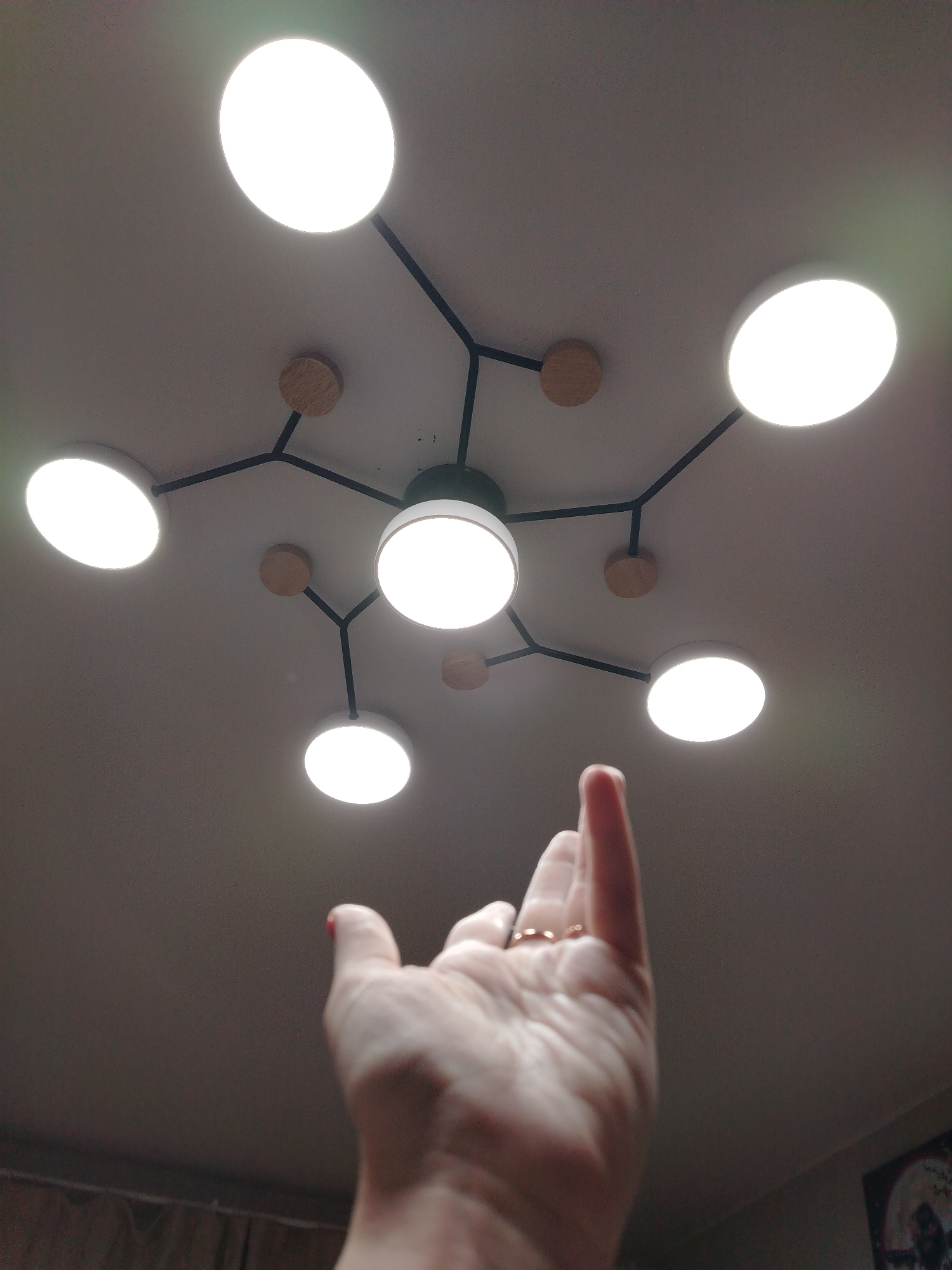 The image captures an upward-looking view of a ceiling light fixture, with a person's hand reaching towards it in an indoor setting, likely in Izhevsk, Russia.
In the foreground, a fair-skinned right hand is raised, palm slightly open and fingers gently curled, extending towards the overhead light. A gold-colored ring is visible on the fourth finger, and the fingertips of some fingers, notably the index and middle, show traces of red nail polish.
The main subject is a modern ceiling light fixture, designed with a central circular light from which multiple thin, black rods branch out. These rods connect to six additional circular light sources, creating a total of seven illuminated white discs. Small, round wooden decorative elements are interspersed along the black rods, adding a contrasting texture and color. All lights are brightly illuminated, casting a strong white glow and producing some minor lens flares in the upper left and lower right of the frame.
The ceiling is a plain, light color. In the lower right corner of the image, a small portion of a framed picture or poster with dark imagery is visible on a wall, suggesting a domestic or commercial interior. The artificial lighting implies it could be evening or night, or simply an interior requiring illumination during the day. No specific weather conditions or visible text are discernible.