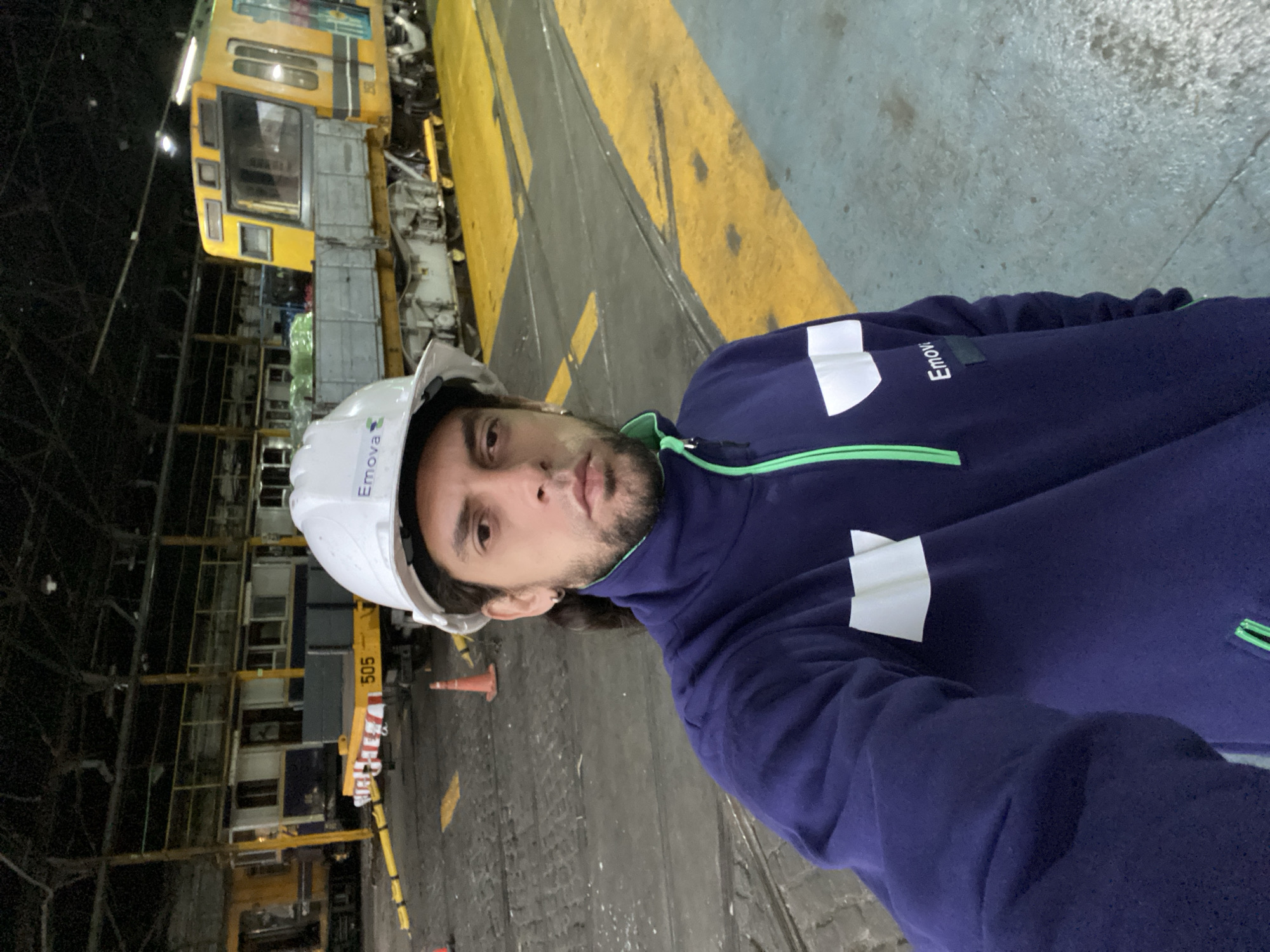 A man with dark hair and a beard stands in a large industrial space, possibly a train maintenance or repair facility. He wears a white hard hat with the "Emova" logo and a blue jacket with a white reflective stripe on the arm. He is facing the camera. Behind him is a yellow train car with the number "505". Yellow safety markings are painted on the floor. The lighting is dim, with artificial lights illuminating the scene. The facility appears to be located in Buenos Aires, Argentina.