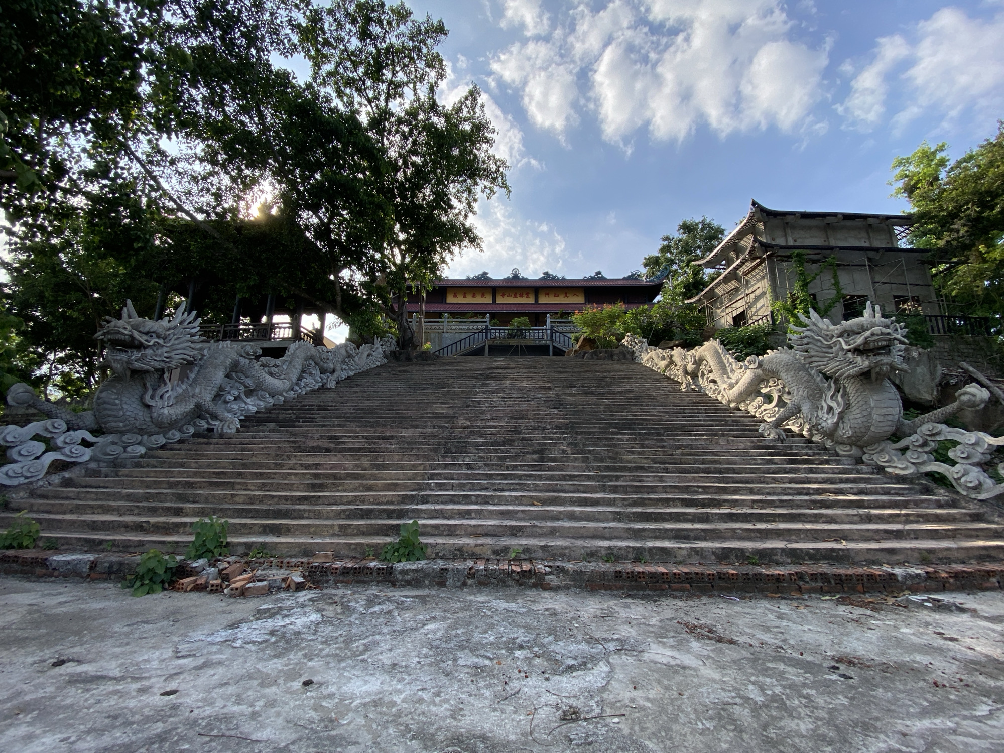 An exterior, low-angle, wide shot captures a grand staircase leading to a temple building. The staircase is flanked by intricately carved stone dragon statues. Trees with dense green foliage are visible on either side of the staircase, with sunlight filtering through the leaves. The temple building, with its distinctive tiered roof and yellow accents, is situated at the top of the stairs. To the right of the temple, a partially constructed building is visible, covered in scaffolding and vine growth. The sky above is blue with scattered white clouds, suggesting a clear day. The base of the staircase shows a concrete ground with scattered bricks and some vegetation growing between the steps. This scene is located in Tay Nha Trang Ward, Vietnam.
