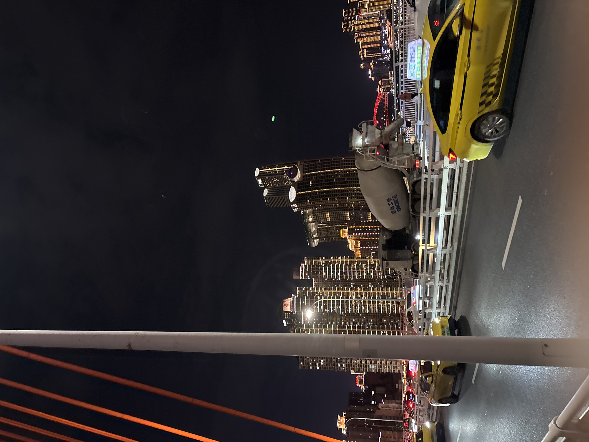 This media file shows a nighttime view of a city skyline in Yuzhong District, China. The scene is dominated by tall, illuminated buildings and a bridge. In the foreground, a white pipe runs horizontally across the frame, with orange cables beneath it.
Several objects are present on the bridge, including a cement mixer truck and at least two yellow taxis. One taxi is clearly visible in the right half of the frame, with its door open and a person inside. The taxi has a checkerboard pattern on its side and text on its roof sign. The other taxi is partially visible in the lower right corner.
The city skyline is characterized by numerous skyscrapers adorned with bright lights, creating a vibrant urban landscape against the dark night sky. The lighting suggests the time of day is night. The presence of the bridge, taxis, and construction equipment implies an active urban environment. No specific weather conditions are apparent, but the clear sky suggests fair weather. The overall impression is one of a bustling city at night.
Visible text includes Chinese characters on the taxi's roof sign and on the side of the cement mixer truck. The taxi also has a checkered pattern on its side and some text on its rear.