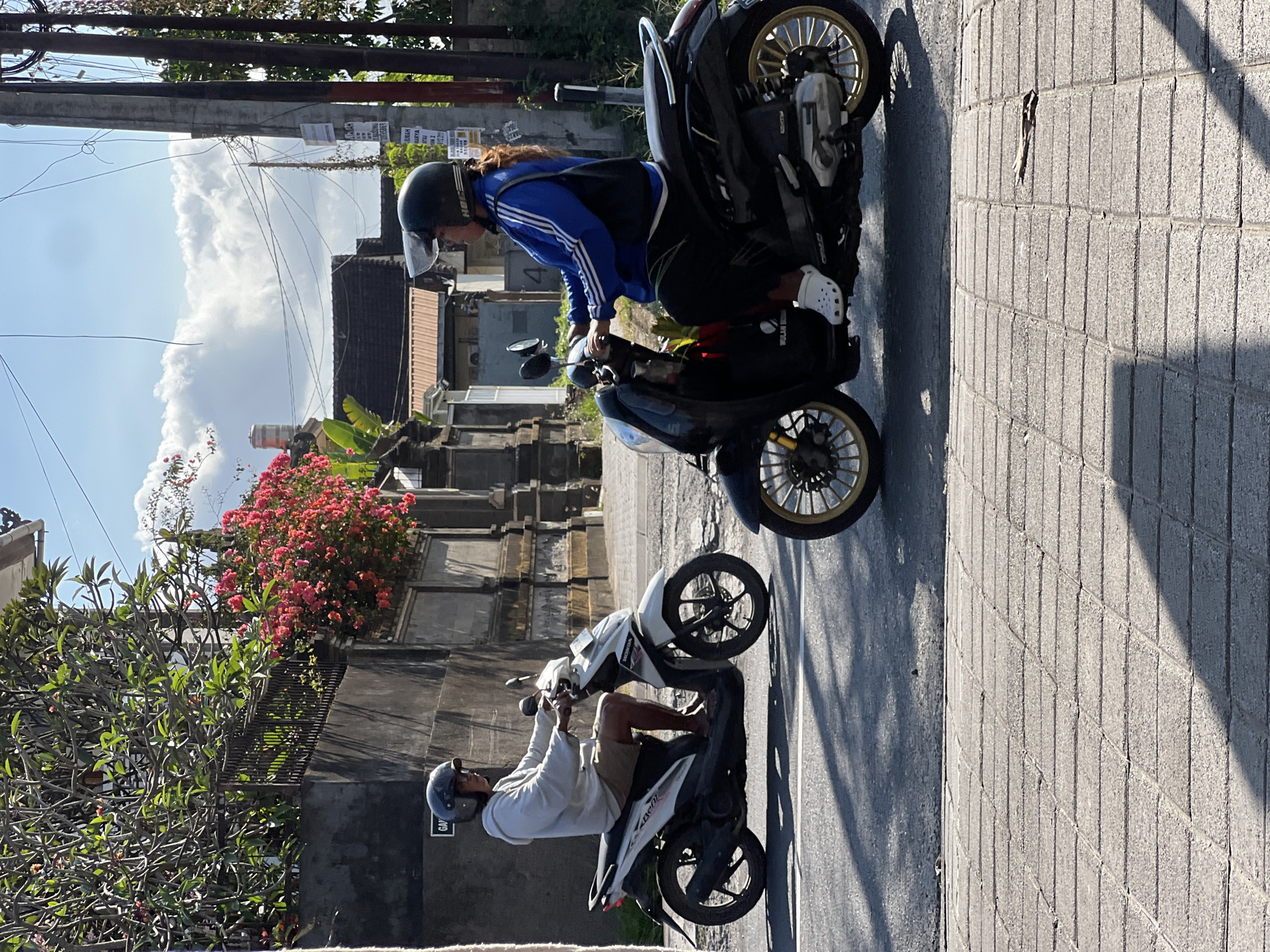 Two people are riding motorbikes on a sunny day in Denpasar, Indonesia. A man in a white hoodie and brown shorts, wearing a black helmet, rides a white scooter on the left. A woman in a blue jacket with white stripes, black pants, and white clogs, also wearing a black helmet, rides a black scooter on the right. They are on a paved road with a sidewalk made of stone blocks. In the background, there are stone walls, lush green trees, and pink bougainvillea flowers. Power lines and utility poles are visible against a bright blue sky with scattered white clouds. The scene appears to be in a residential or semi-urban area.