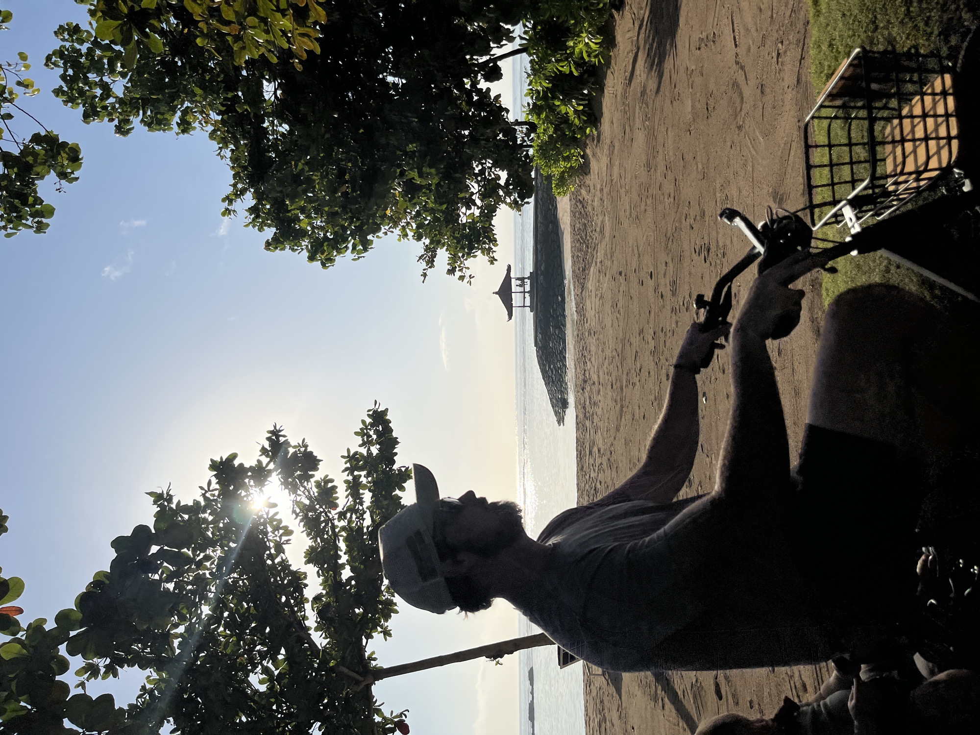 In Kuta Selatan, Indonesia, a man with a beard and a baseball cap is silhouetted on a bicycle on a sandy beach, facing the ocean. The sun shines brightly behind him, creating lens flare through the leaves of a tree. In the distance, a small pavilion sits on a rocky outcrop in the water, with two figures visible underneath. The beach is sparsely populated, with footprints in the sand. Green bushes and trees frame the scene on either side, with the ocean stretching out to the horizon under a clear blue sky. A basket is attached to the front of the bicycle.