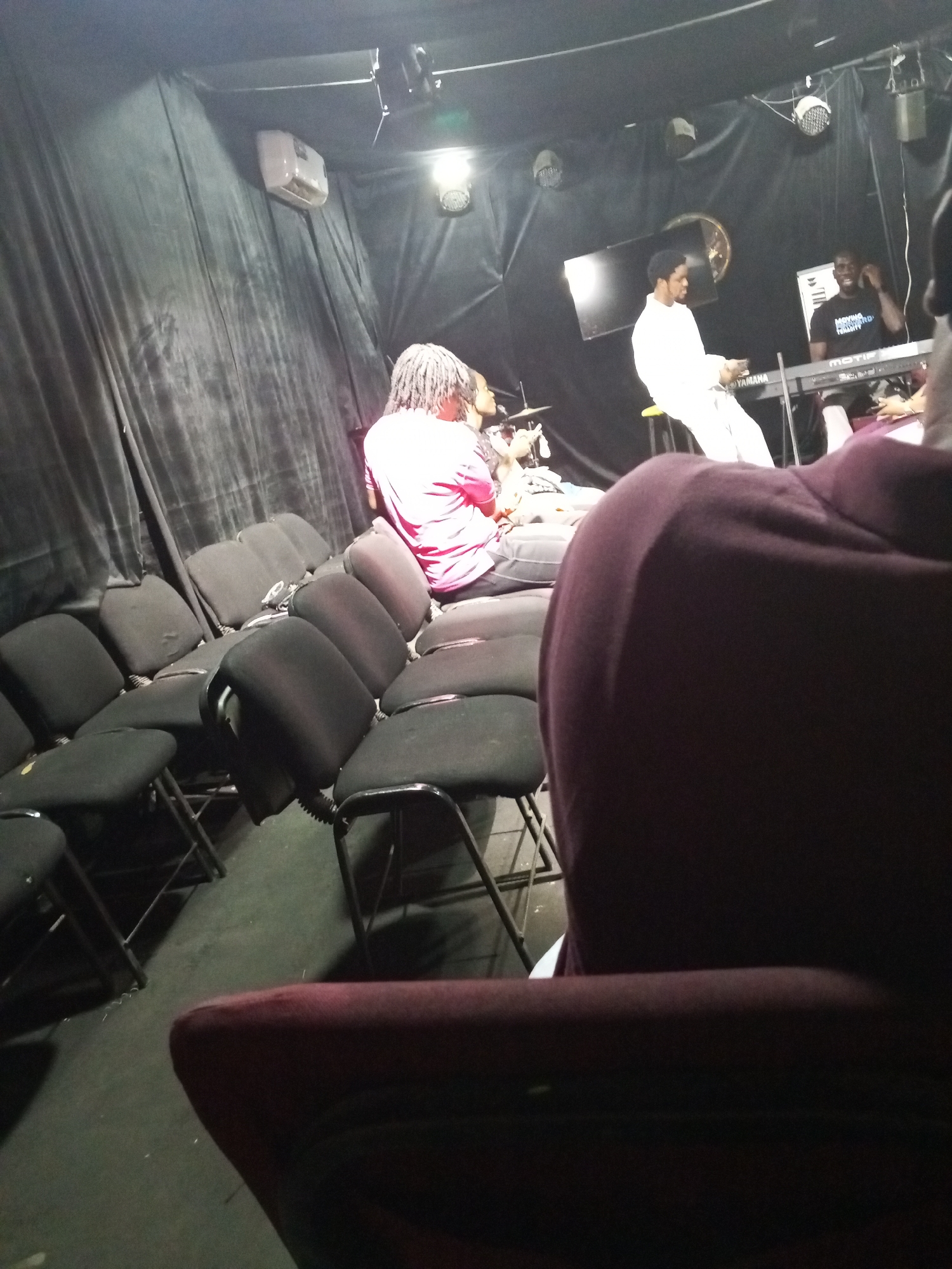 The image shows a music performance inside a dark room or studio. Three individuals are on stage. One, wearing a red shirt and dreadlocks, is seated behind a drum set. Another individual, dressed in white, is playing a Yamaha keyboard while seated on a stool. To his right, another person wearing a black t-shirt with white writing, possibly "Moving Forward Initiative," is also present behind the keyboard. The stage area is set against black draping. Several empty black folding chairs are positioned in the foreground, indicating an audience area. A large portion of the foreground is occupied by a blurred object (possibly a chair or part of a person), creating a sense of being in the audience. The environment is dimly lit with stage lighting, including spotlight fixtures on the ceiling. The location is determined to be in Abuja, Nigeria, although specific details within the image do not directly confirm this, which is only from external instruction.