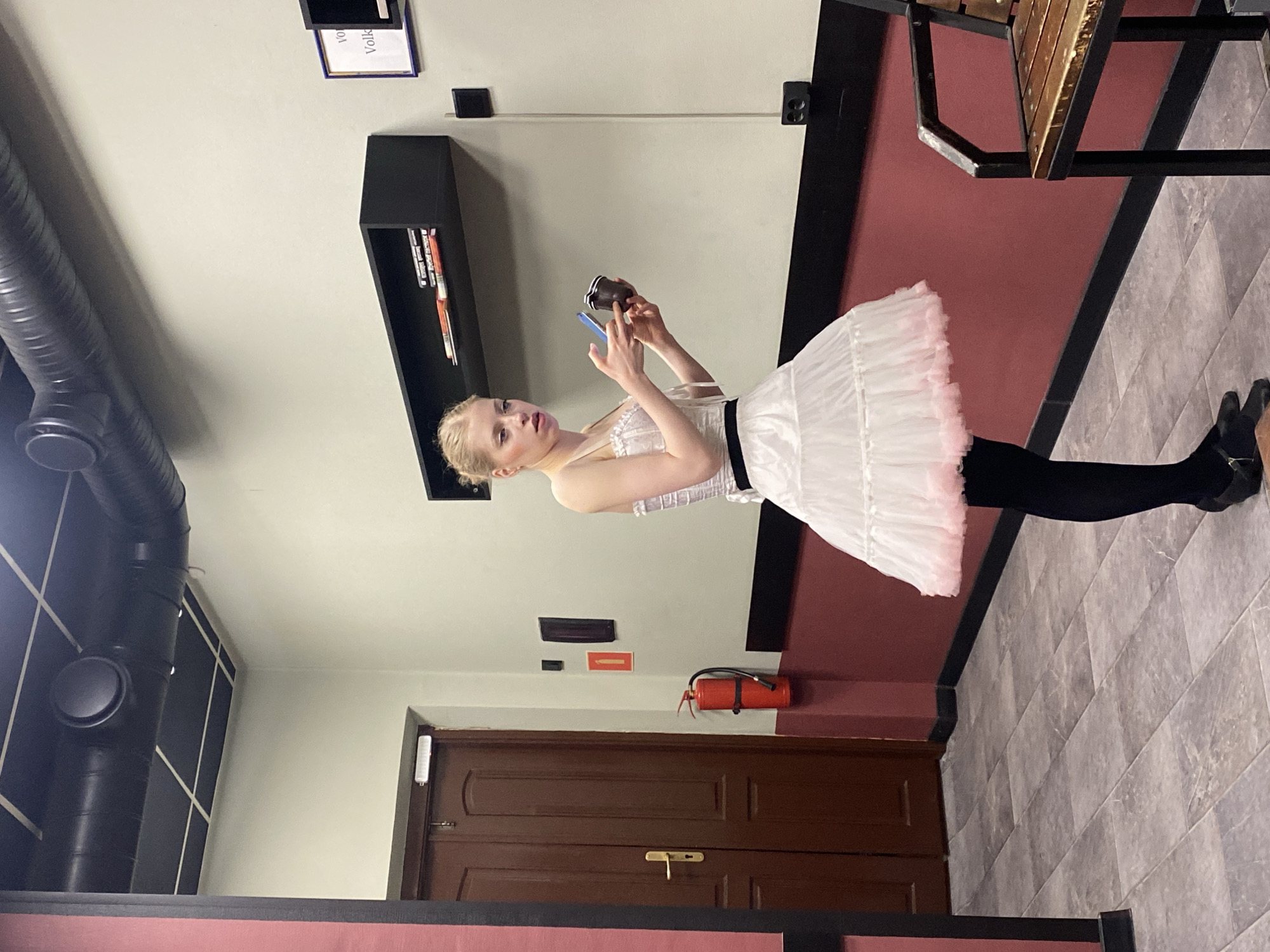 The image shows a fair-skinned young woman with blonde hair, dressed in a white tutu, black tights, and black ballet shoes. She is holding a blue smartphone and a brown object (possibly a cup) in her hands, looking at them with a focused expression.
The background suggests an indoor space, likely a dressing room or backstage area. A dark brown door is visible in the lower part of the frame, with a fire extinguisher mounted on the wall above it. There's also a black shelf with books, and a framed paper with "Volk" printed on it. The floor is covered in gray tiles. In the top left corner of the image, there is a black ventilation system visible. A dark red horizontal element appears in the lower part of the frame, possibly part of a wall or furniture. There is some wooden furniture visible in the upper right of the image.
No information is provided regarding the city and the location of the image.