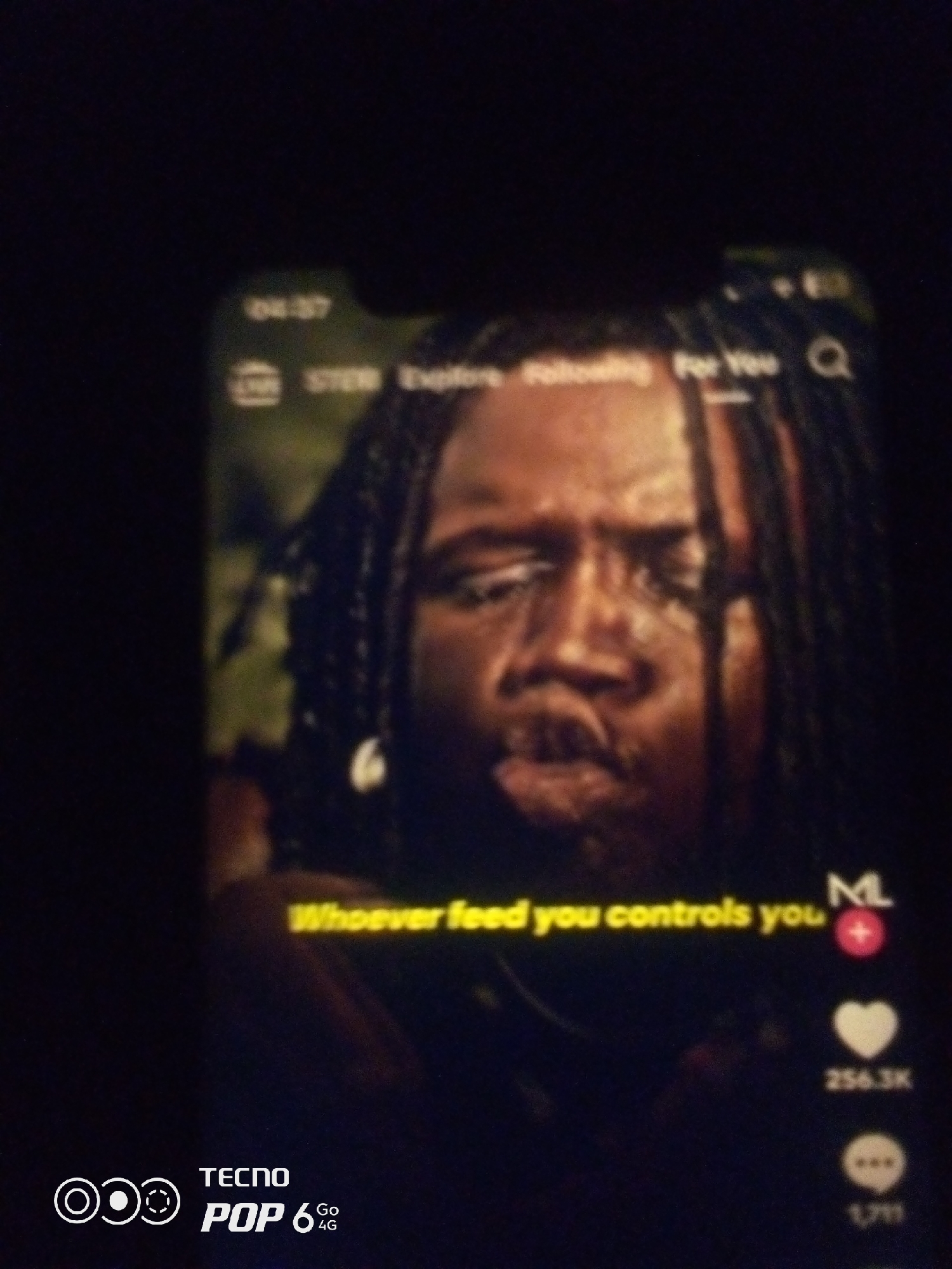 The image shows a close-up of a smartphone screen displaying a video. The video features a man with braided hair, looking intently at the camera. Text overlaid on the video reads "Whoever feed you controls you." The smartphone also displays various app interface elements, including a timestamp "04:37," navigation options like "LIVE," "STER," "Explore," "Following," and "For You," a search icon, and social media interaction icons such as a heart, a comment bubble, and a share icon with a count of "256.3K" likes and "1,711" comments. The lower left corner of the screen shows the "TECNO POP 6 Go 4G" branding. The background of the video appears to be a blurry, natural environment. There is no information within the image that specifies a location in Howayi, Nigeria.